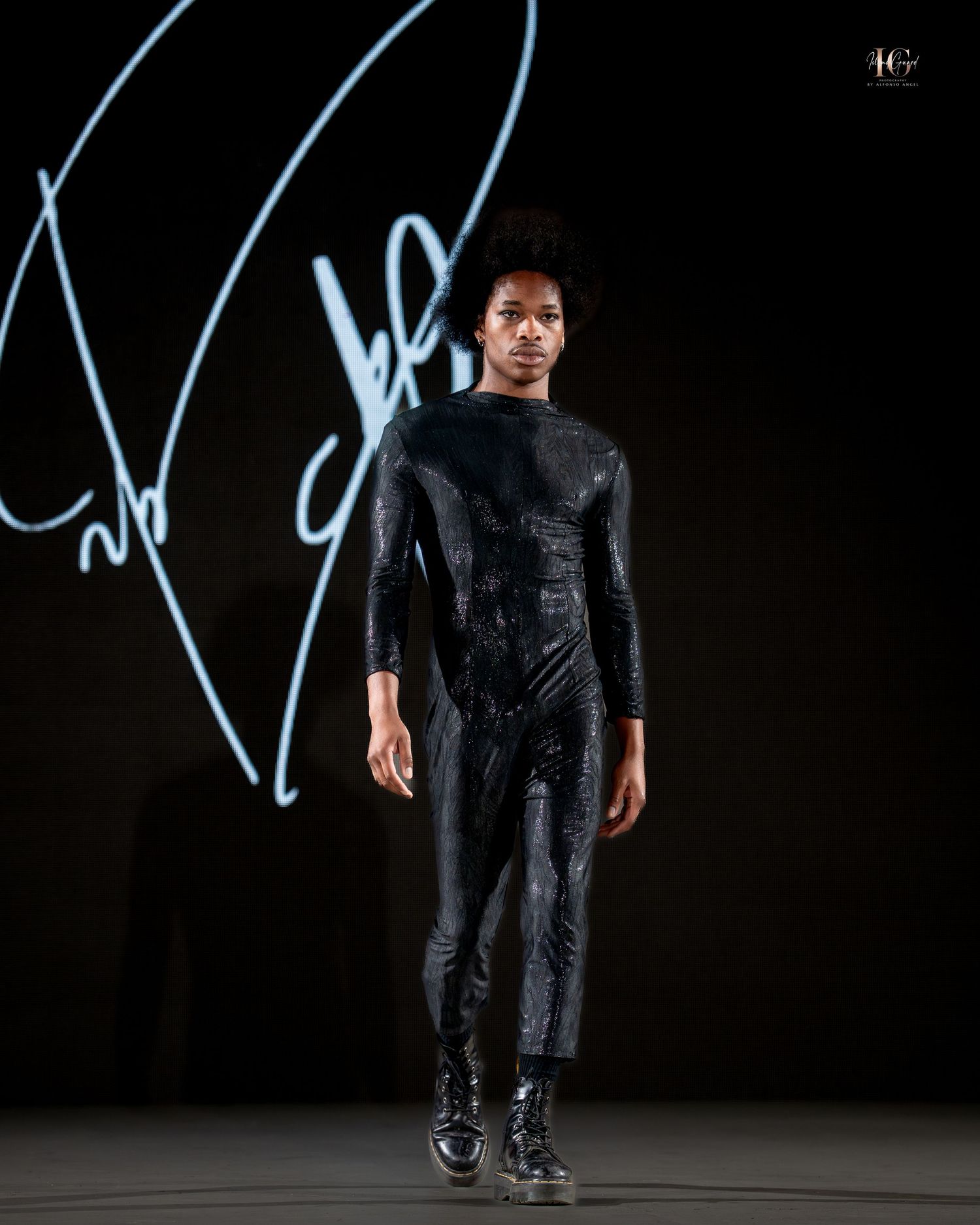 Figure in a shiny black outfit performs on a dark stage with neon blue lighting effects in the background.