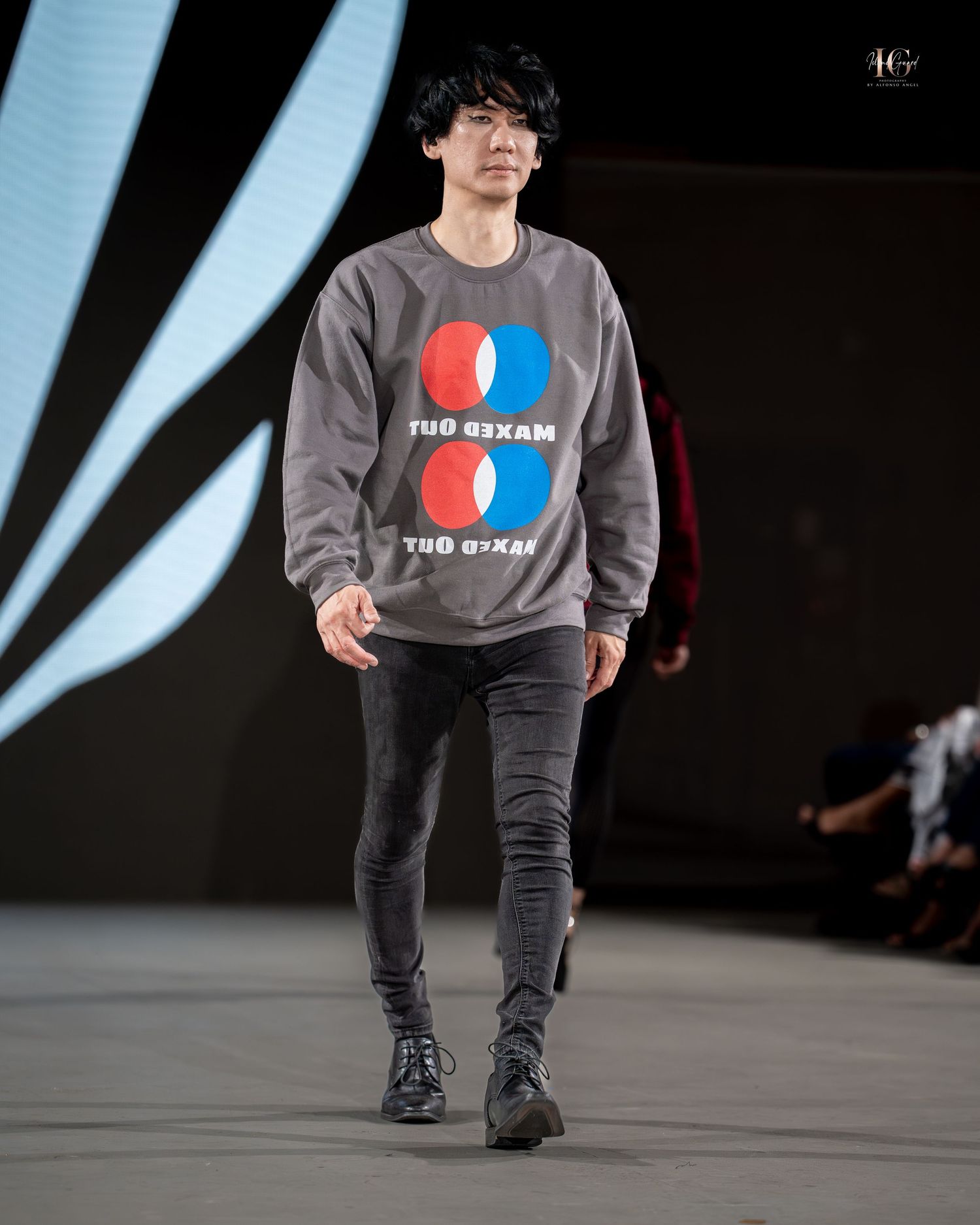 Model walks runway wearing grey sweatshirt with graphic logo and dark pants during fashion show.