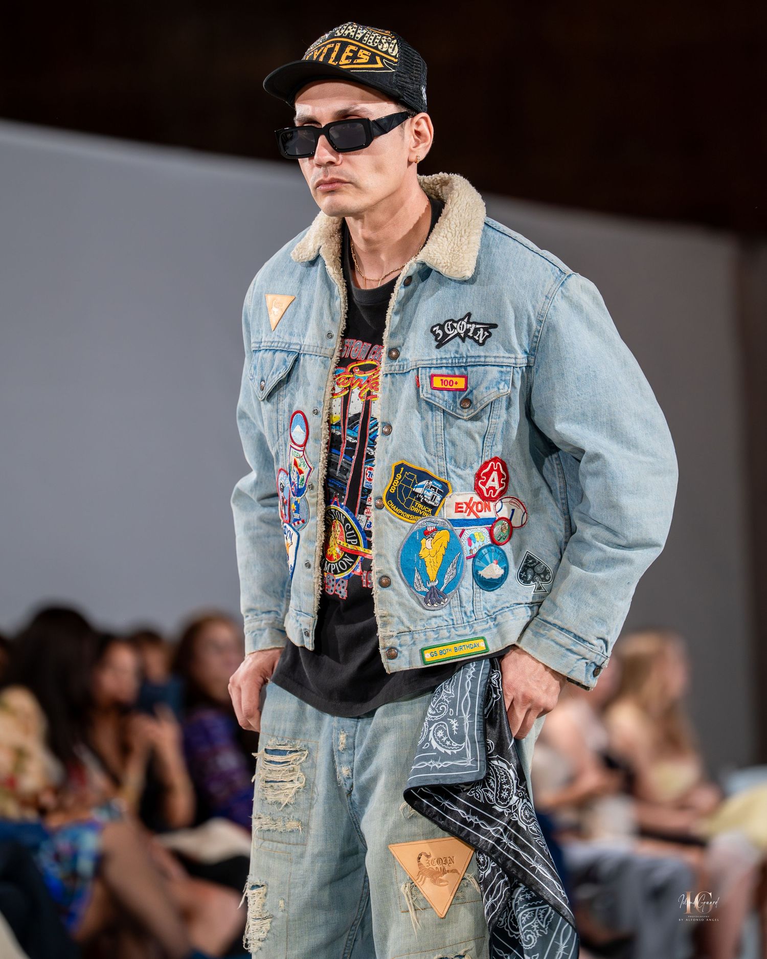 Model walks runway wearing denim jacket with patches and pins during fashion show.