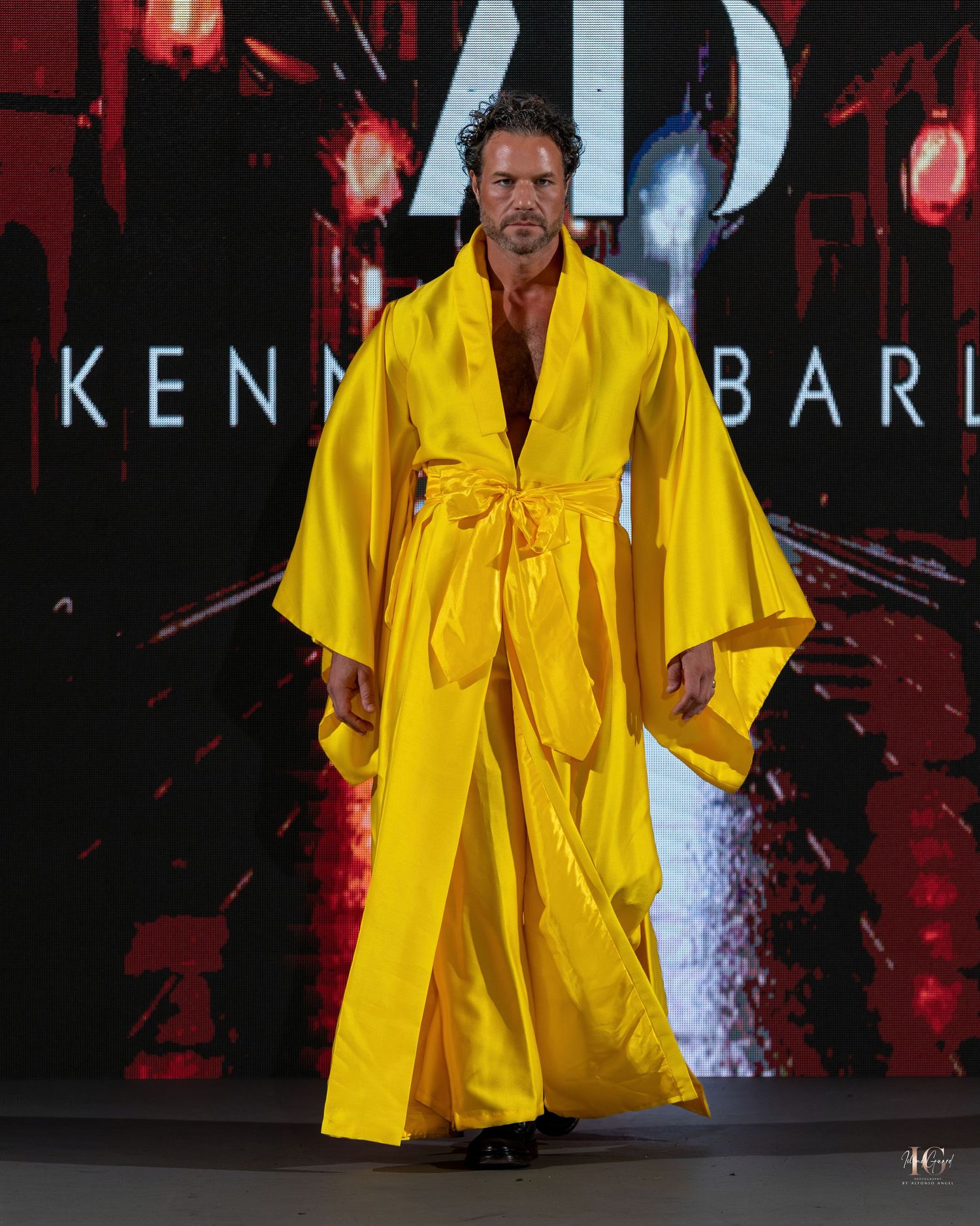 A model walks the runway wearing a bright yellow silk kimono-style robe with a matching belt at a fashion show.
