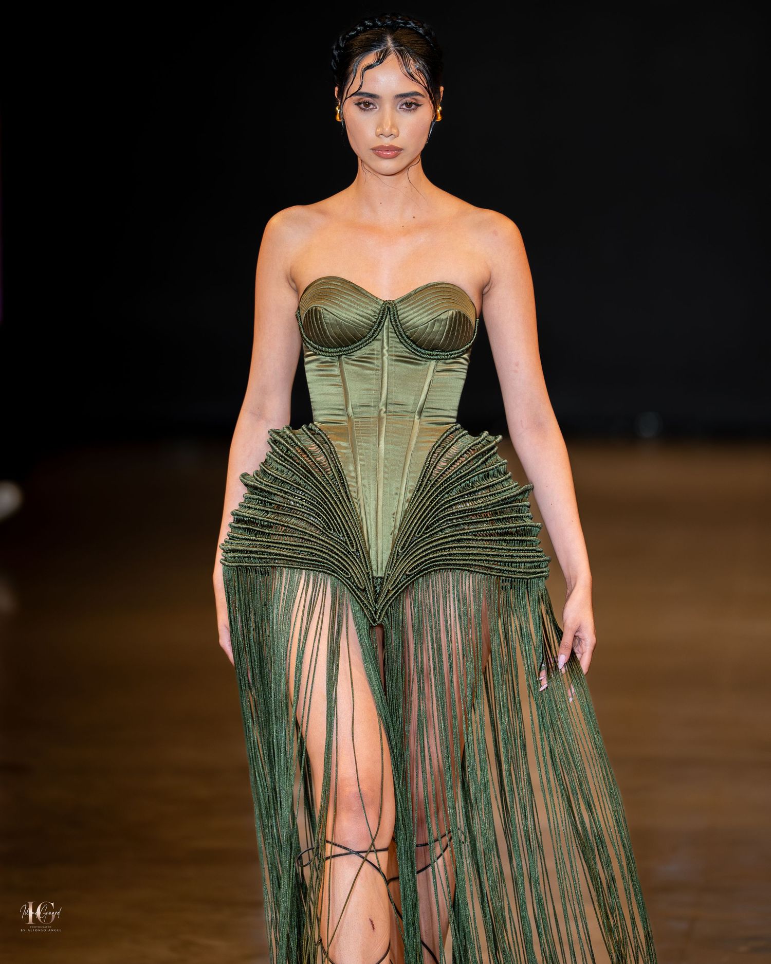 A model walks the runway in an olive green corset dress with dramatic pleated sheer panels at a fashion show.