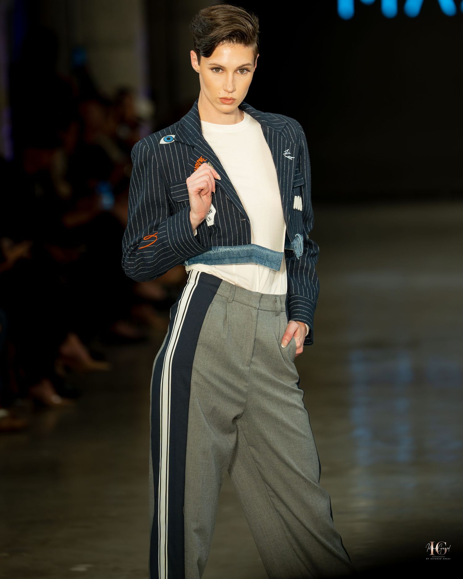 Fashion model walks runway in pinstriped blazer with cream turtleneck and grey pants with white side stripes.