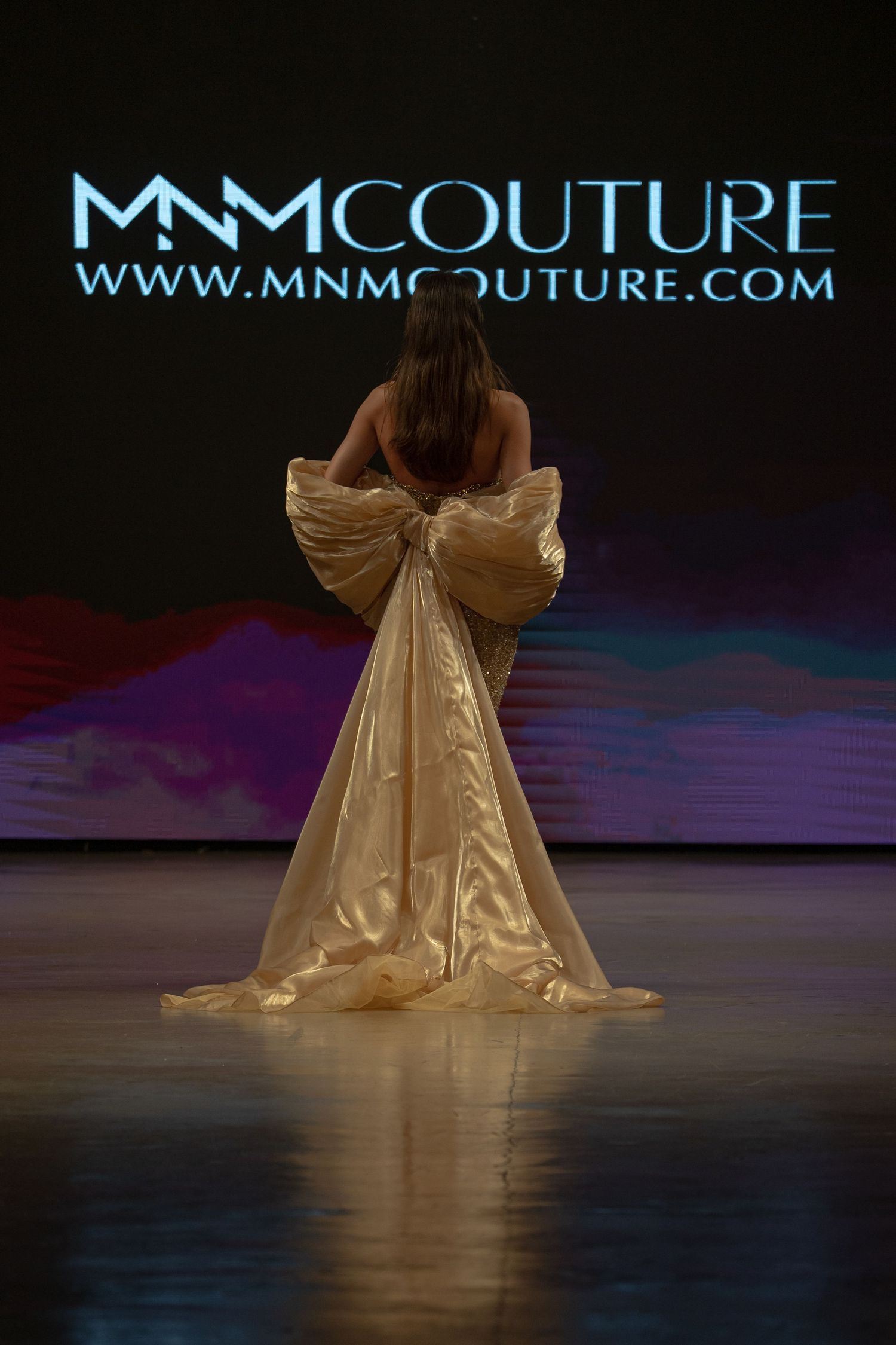 A golden evening gown with dramatic ruffles flows across a dark stage during a fashion show presentation.