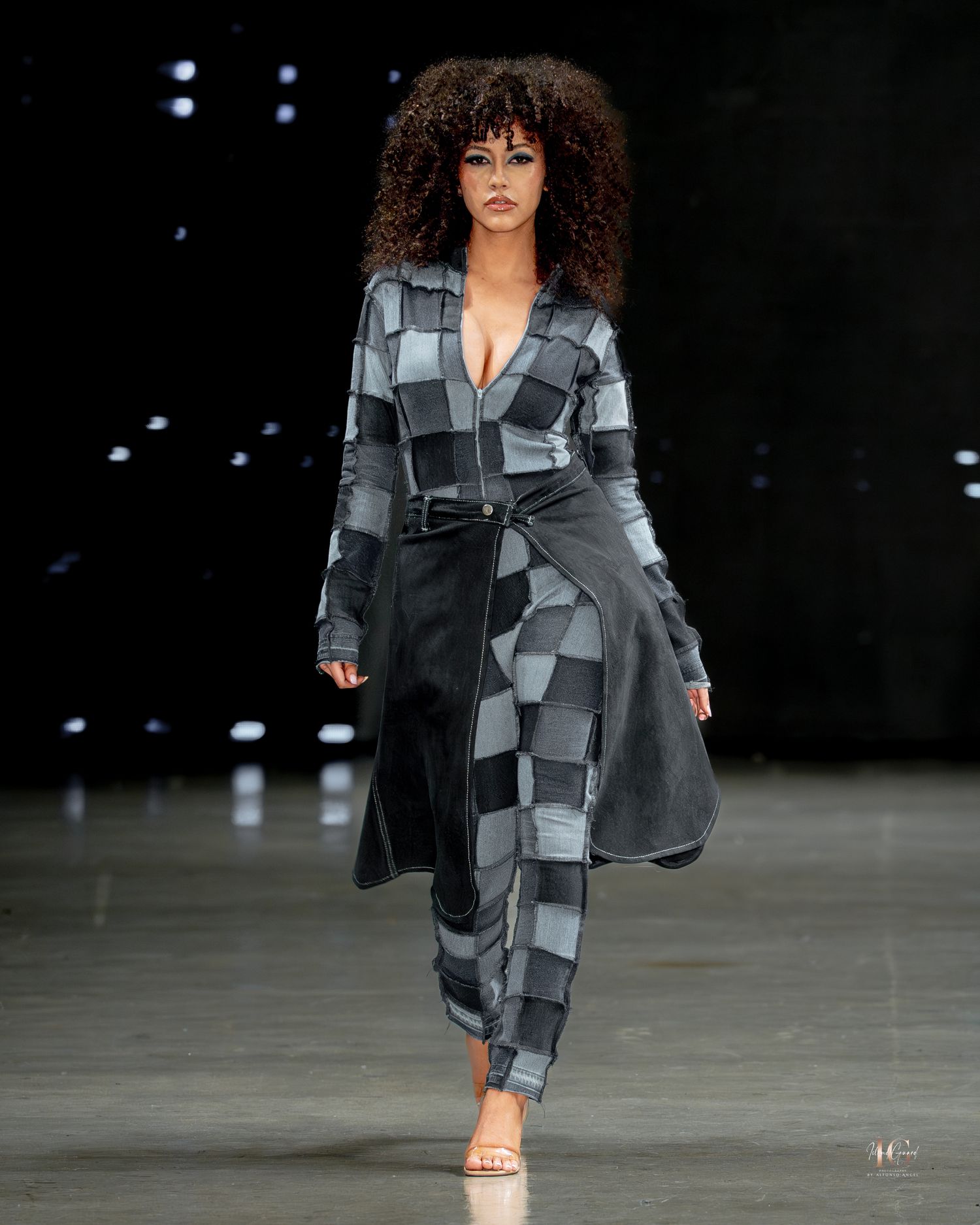 Model walks runway in grey and black geometric patchwork coat and pants ensemble with strappy heels during fashion show.