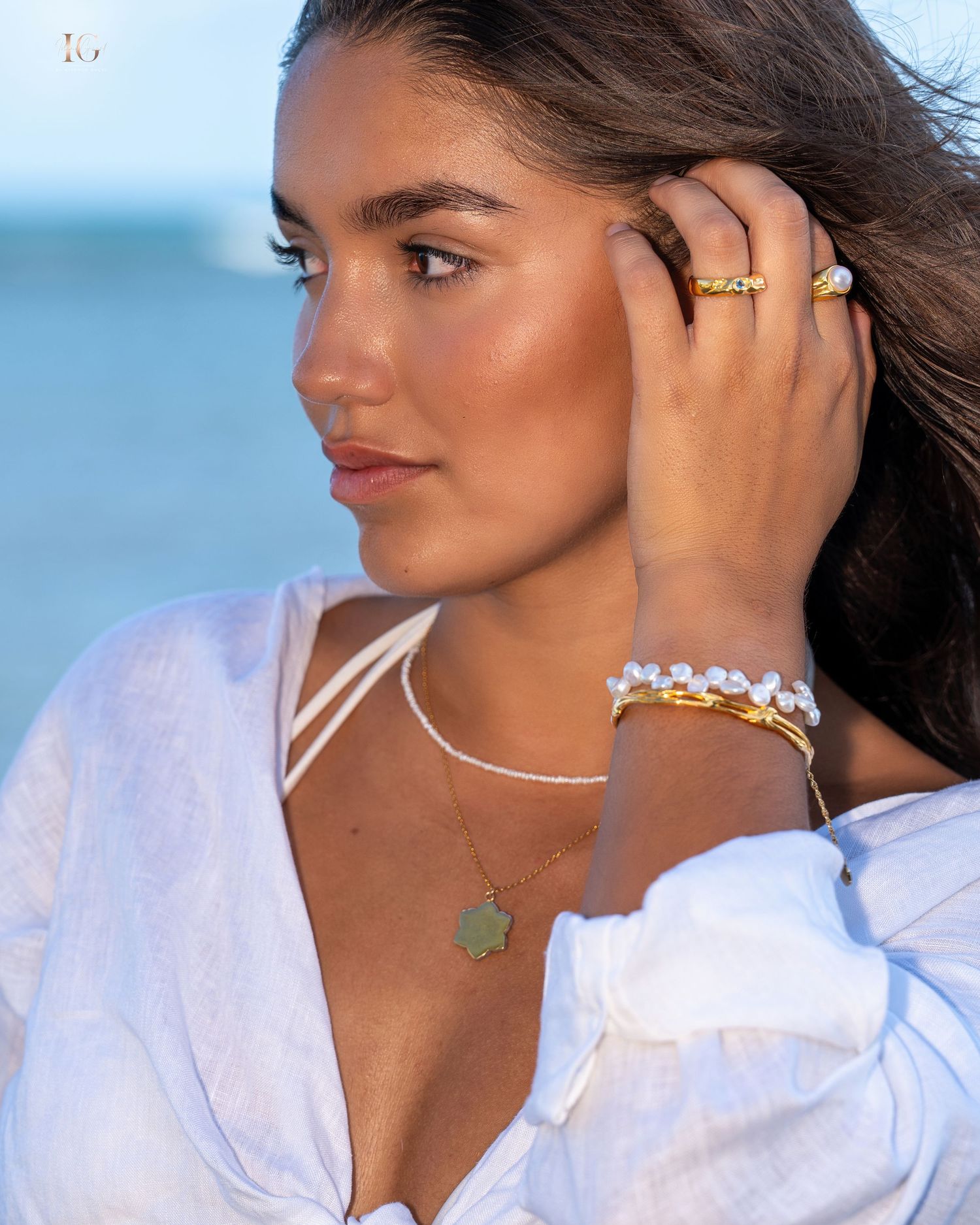 Model wearing a white top and delicate jewelry poses at sunset with an ocean backdrop and windswept hair.