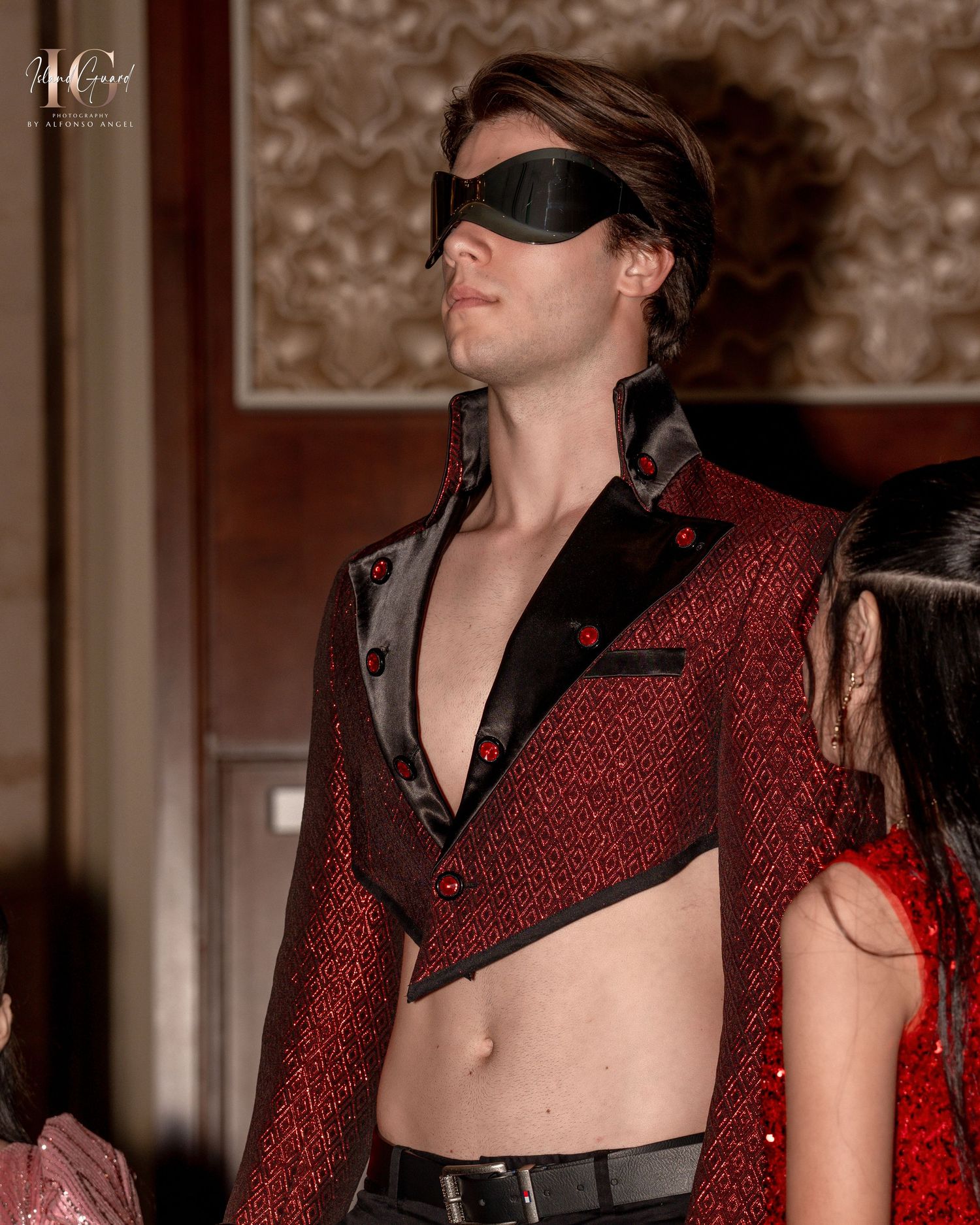 Musicians wearing red and black outfits perform with dramatic lighting against a patterned wallpaper backdrop.