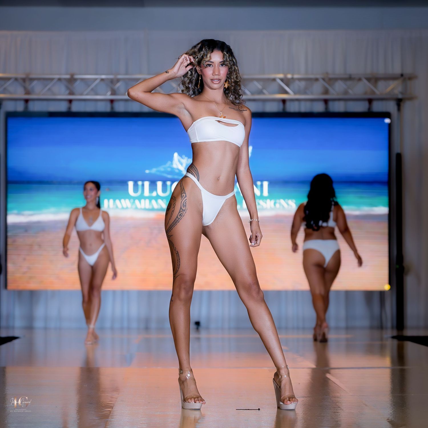 Models present white swimwear collection on beach-themed runway with ocean backdrop.
