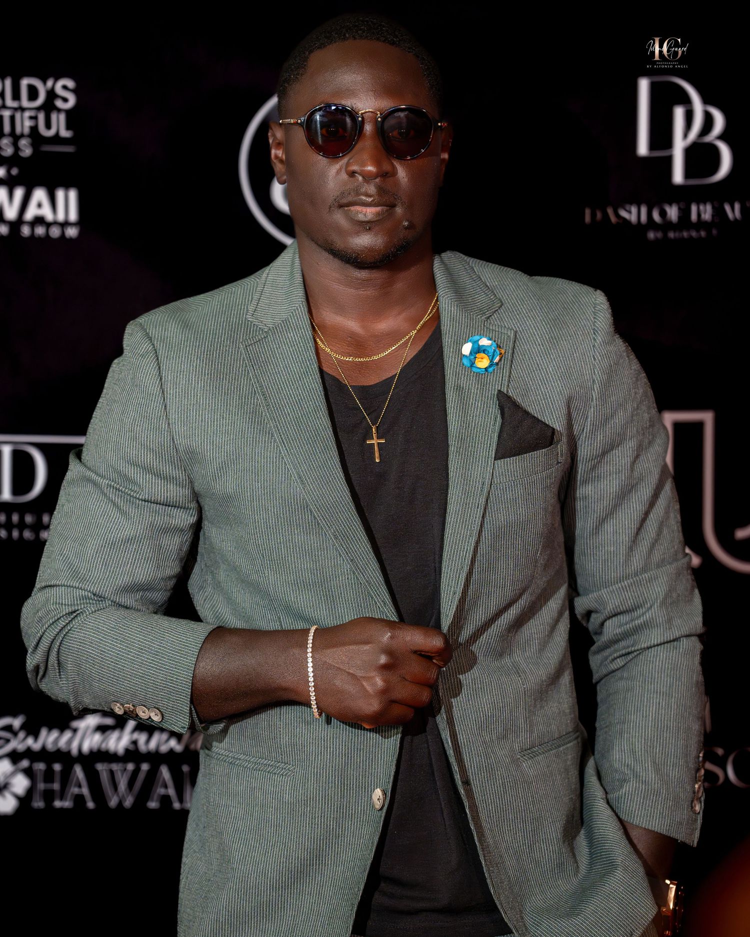 Person in sage green blazer and black t-shirt poses at fashion event wearing sunglasses.