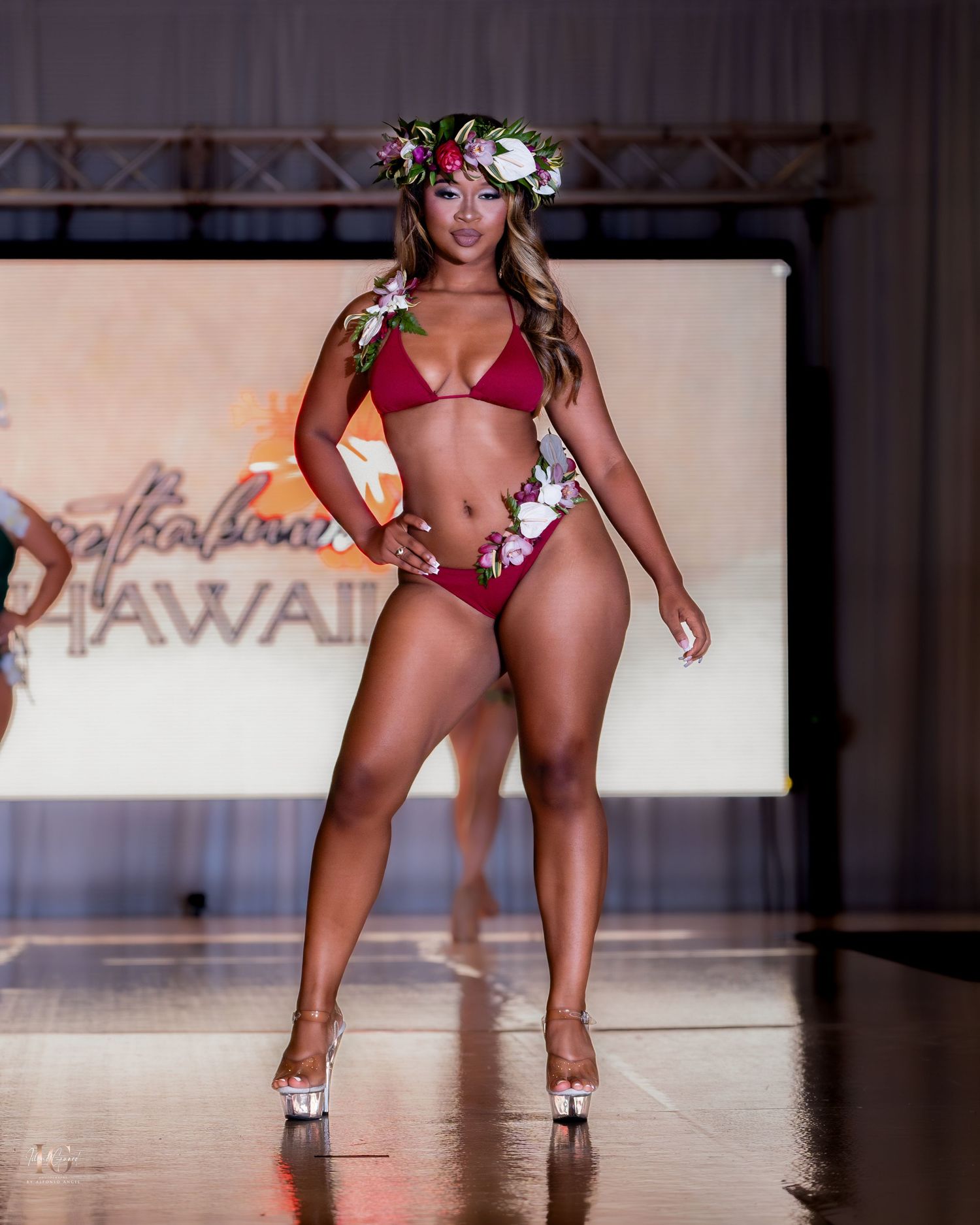 Models wearing floral-accented red swimwear walk runway during tropical-themed fashion show.