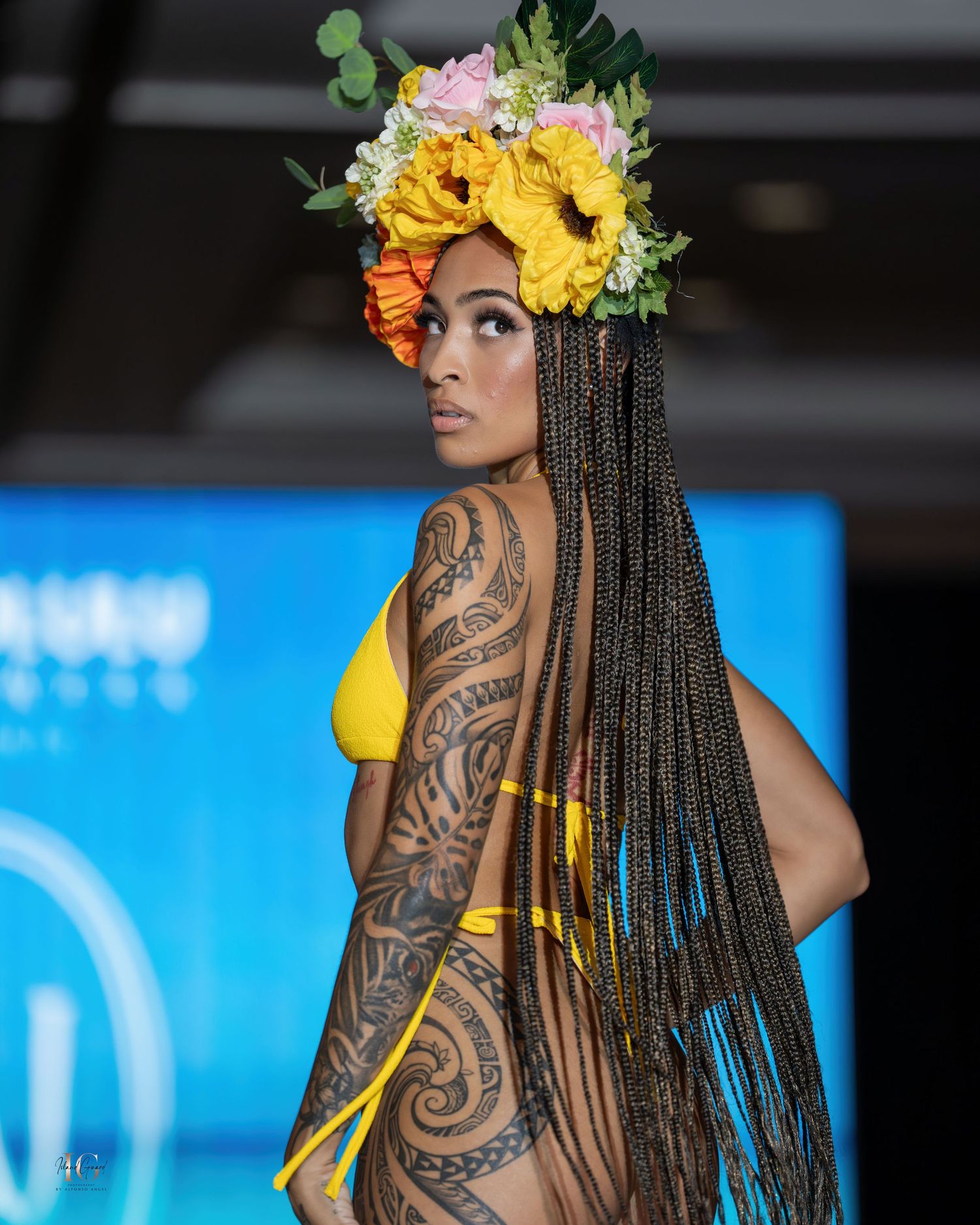 Model showcases intricate tribal tattoos and long braids with yellow floral headpiece at swimwear show.