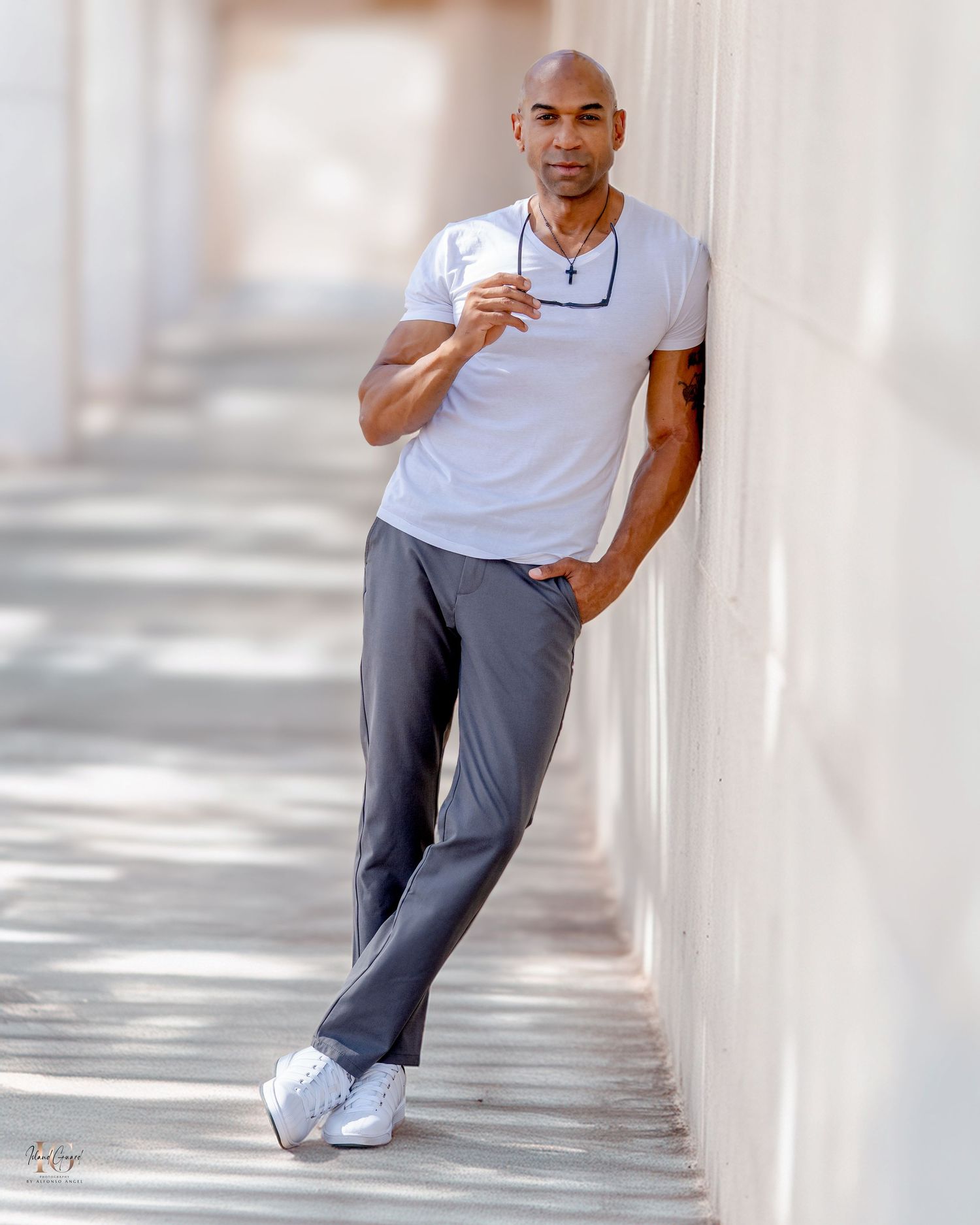Athletic man in white t-shirt and gray pants poses casually against a bright wall in an outdoor hallway.