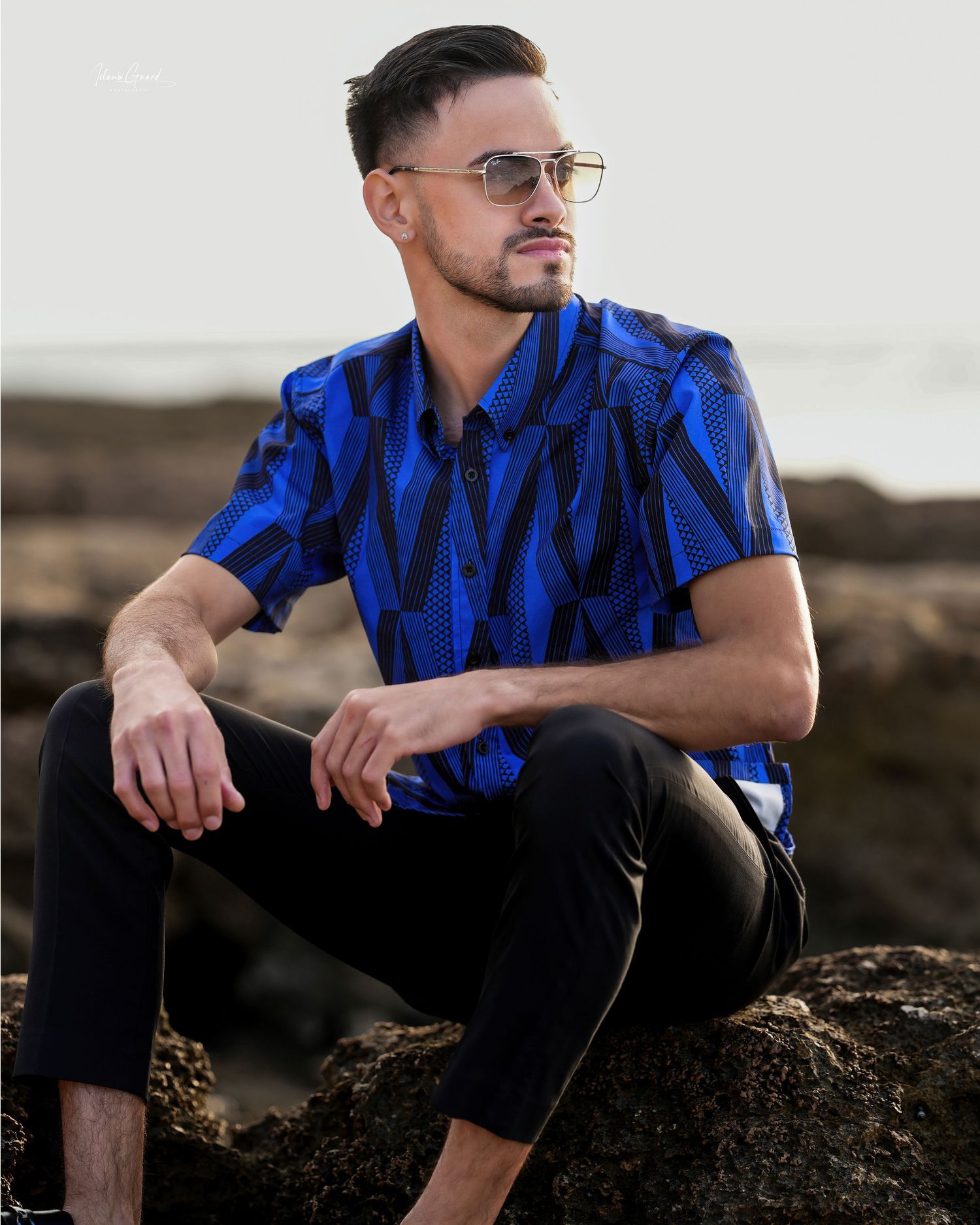Person wearing a vibrant blue patterned shirt and sunglasses sits on coastal rocks at sunset.