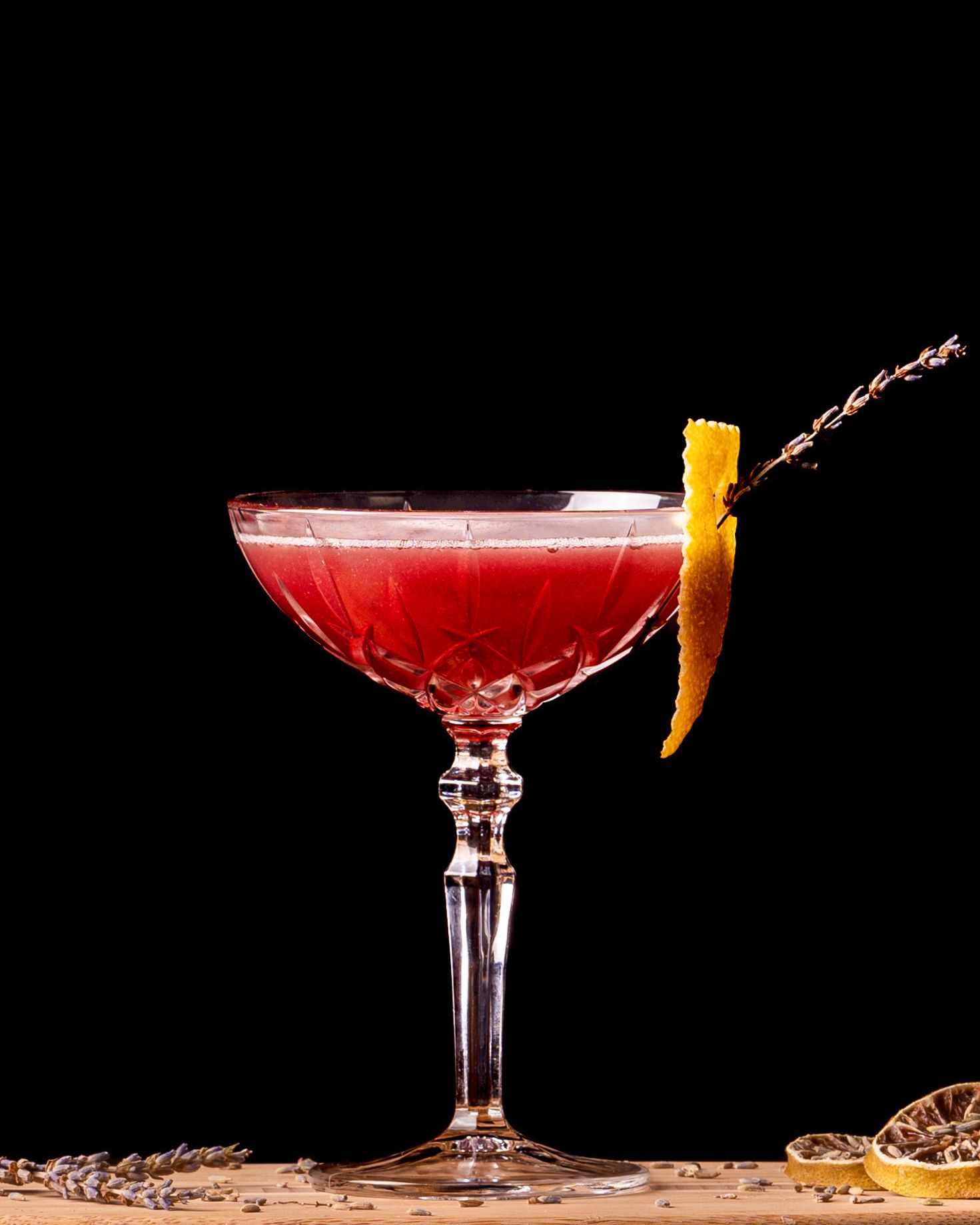Red cocktails in coupe glasses garnished with citrus peels on a dark background.