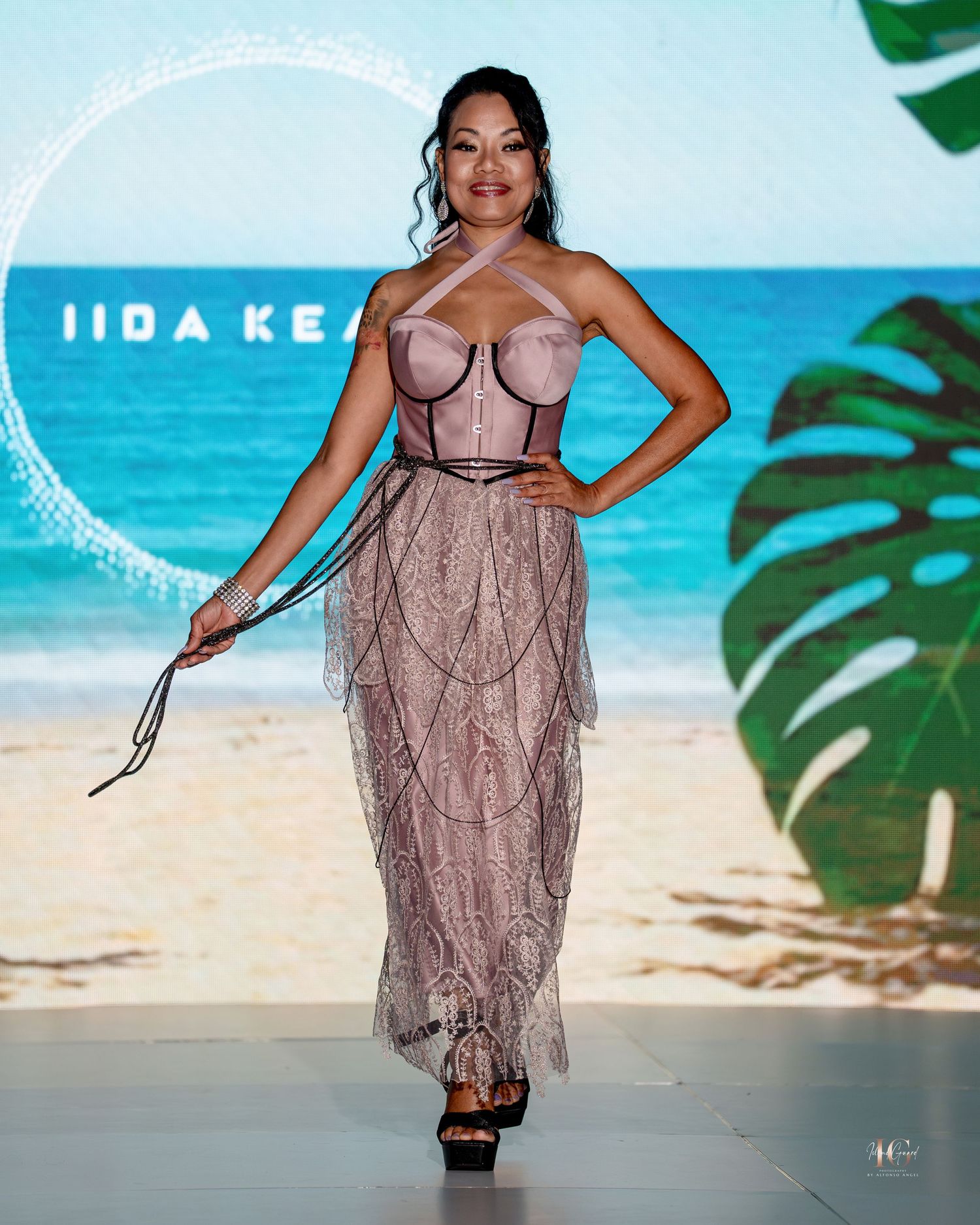 Model walks runway in beige lace lingerie dress at tropical-themed fashion show on beach backdrop.
