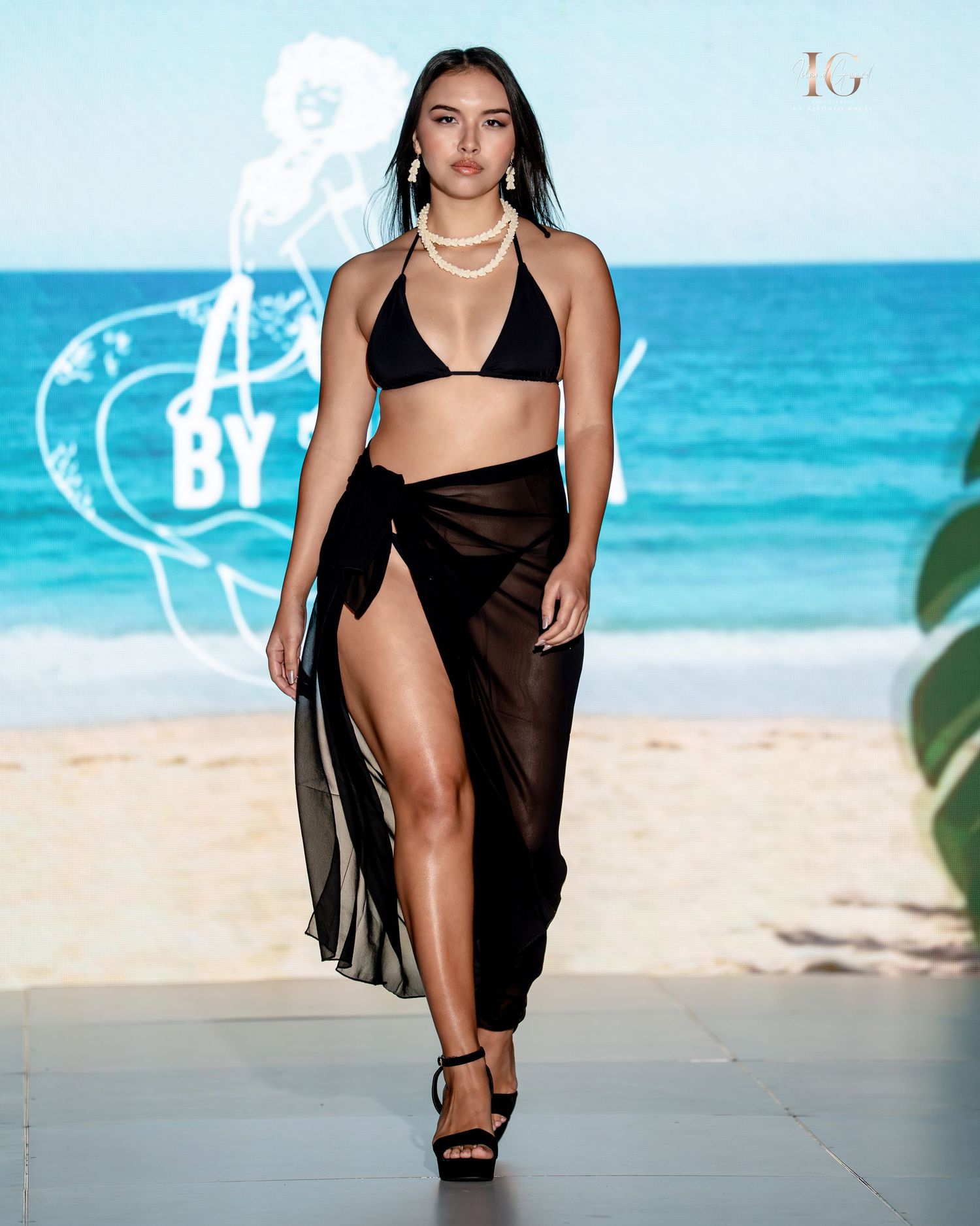 Model in black swimwear and sheer cover-up walks down beachside runway with turquoise ocean backdrop.