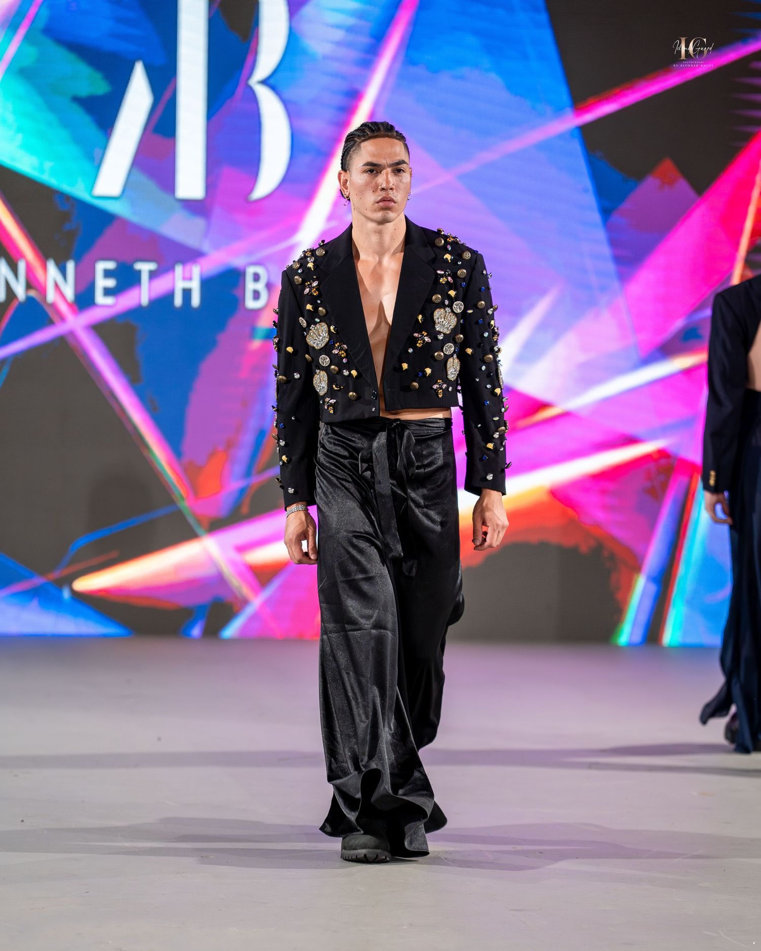 A model walks the runway in a black sequined blazer and pants against a vibrant neon-lit backdrop.