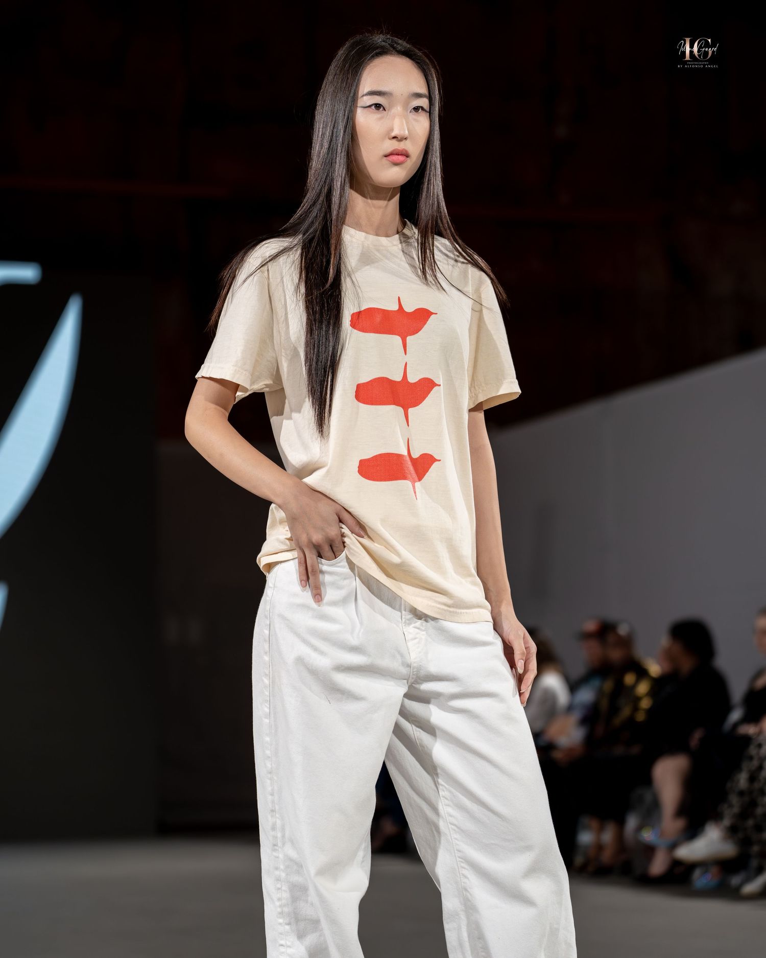 Model walks runway wearing cream t-shirt with red graphic and white pants during fashion show.