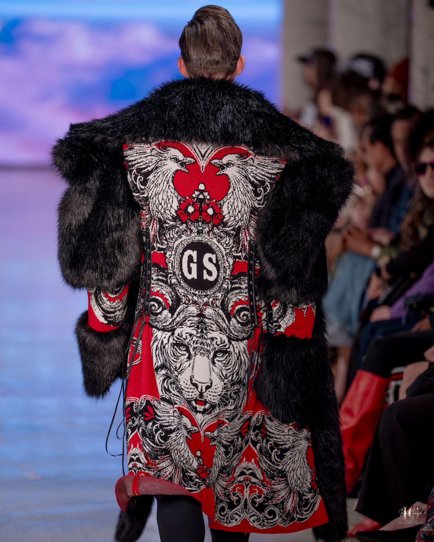 Fashion model on runway wearing black fur jacket with ornate red and white graphic design on back.