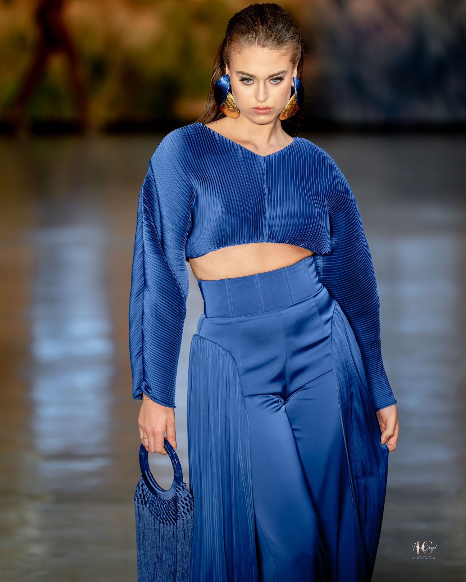 A fashion show model walks the runway in a flowing royal blue asymmetrical dress with pleated details.