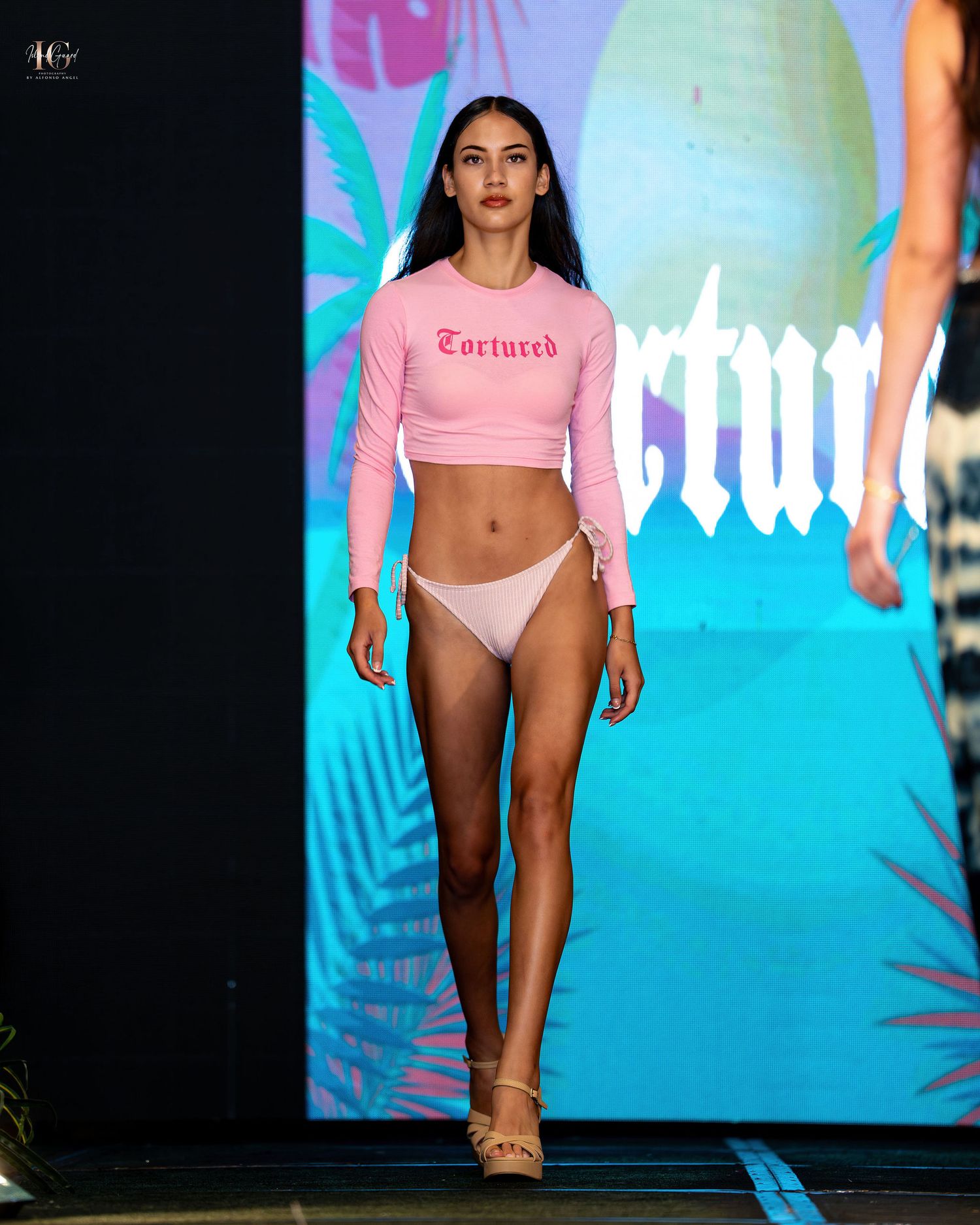 Models walk the runway in pink and white swimwear at a tropical-themed fashion show with blue lighting effects.