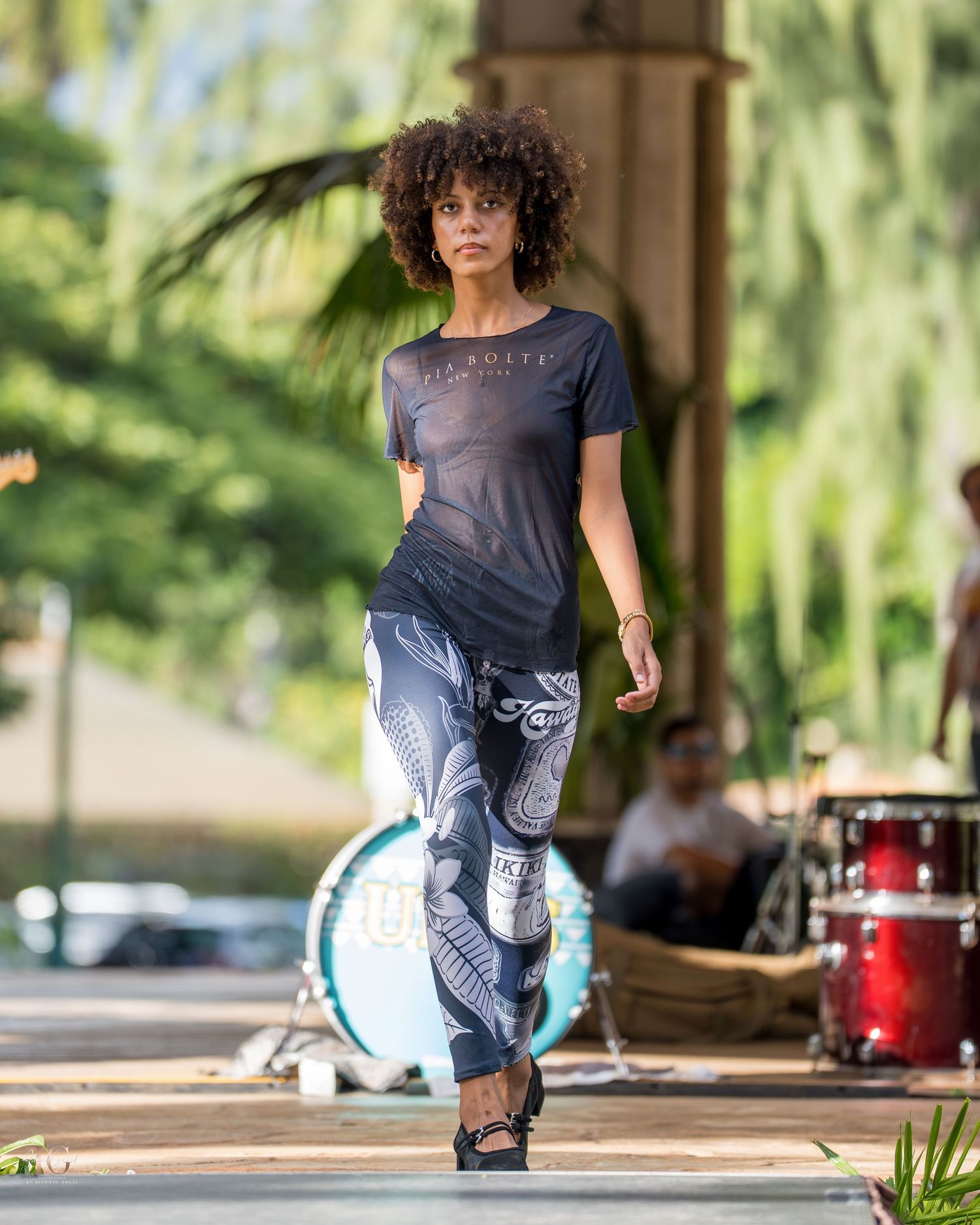 A dancer in flowing clothes performs a graceful movement sequence under a covered outdoor space with palm trees.