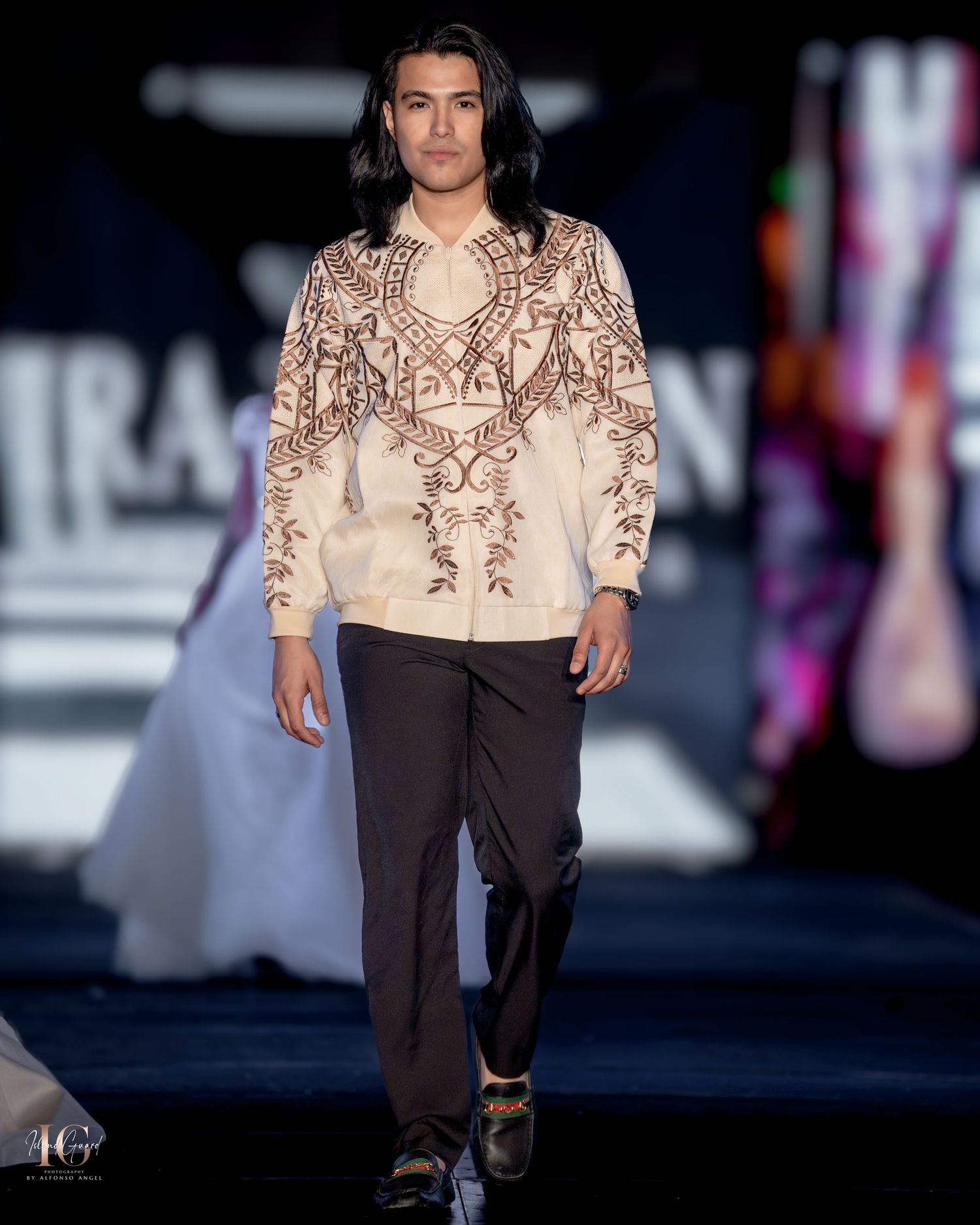Model walks runway in cream embroidered top paired with dark pants against blue lighting.