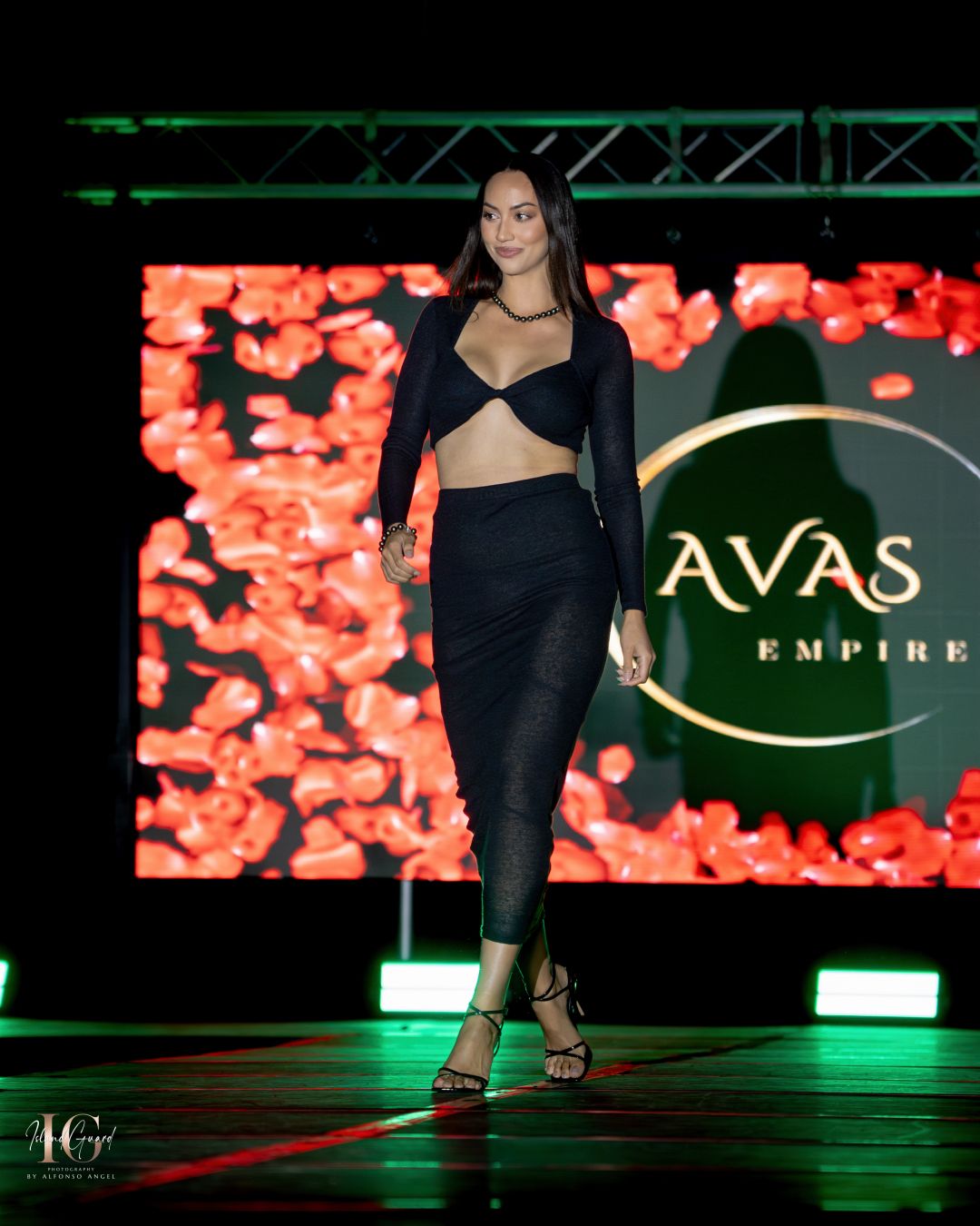 Model walks runway in black cutout ensemble against backdrop with red floral design and AVAS logo.