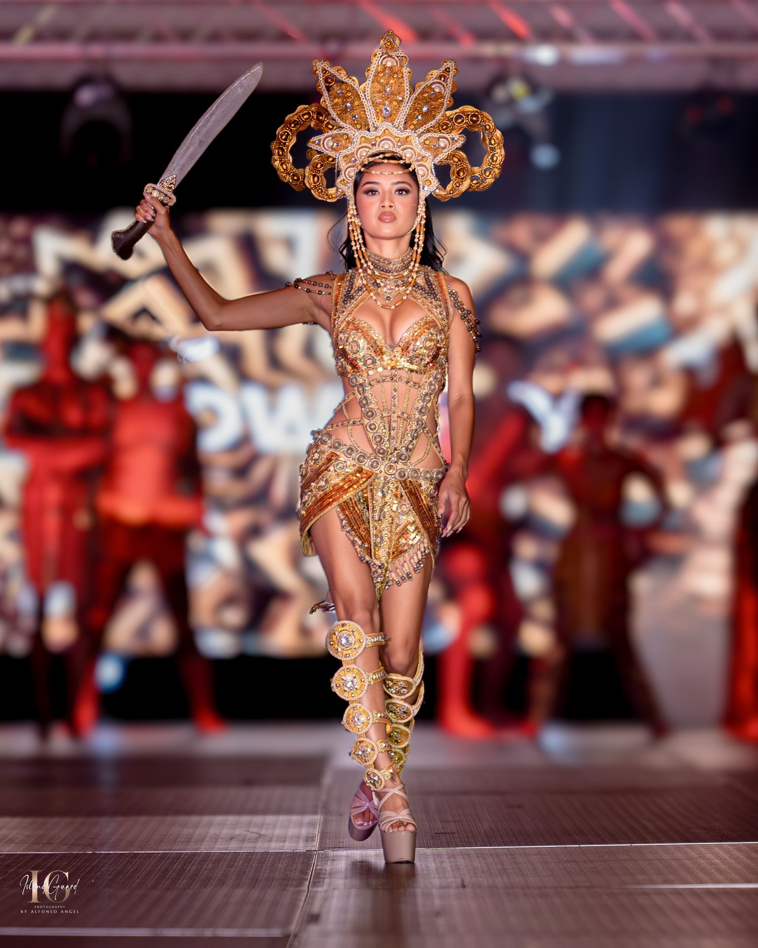 Fashion model in gold warrior-inspired costume with ornate headdress and sword struts down illuminated runway.
