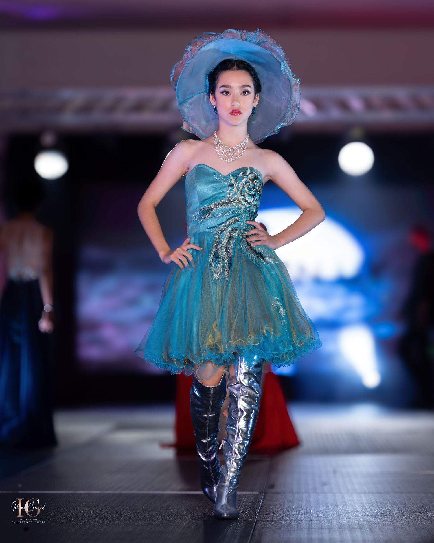 Whimsical teal party dress with matching hat and metallic boots on fashion runway.
