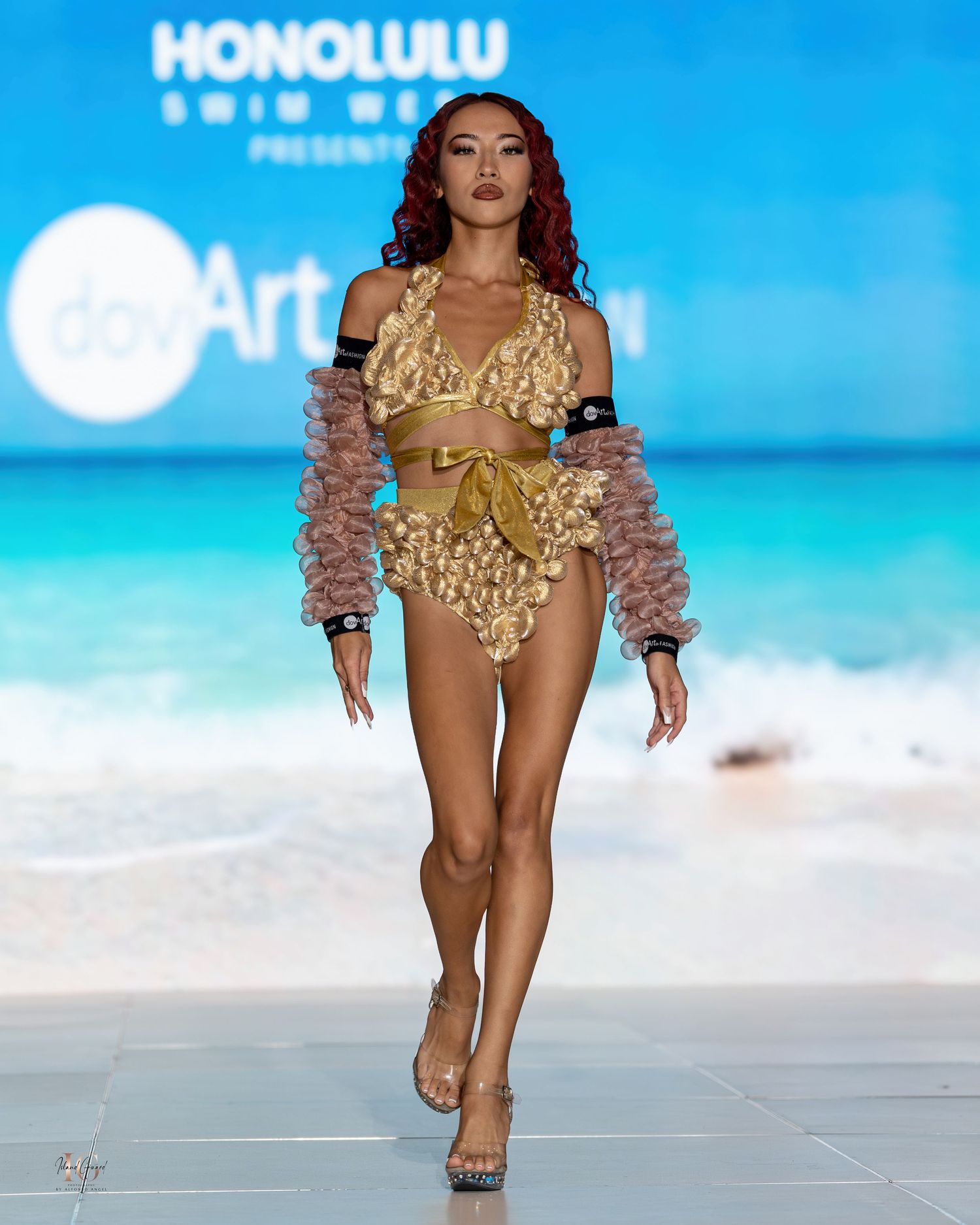 Gold bikini with ruched details modeled on tropical runway with beach background.