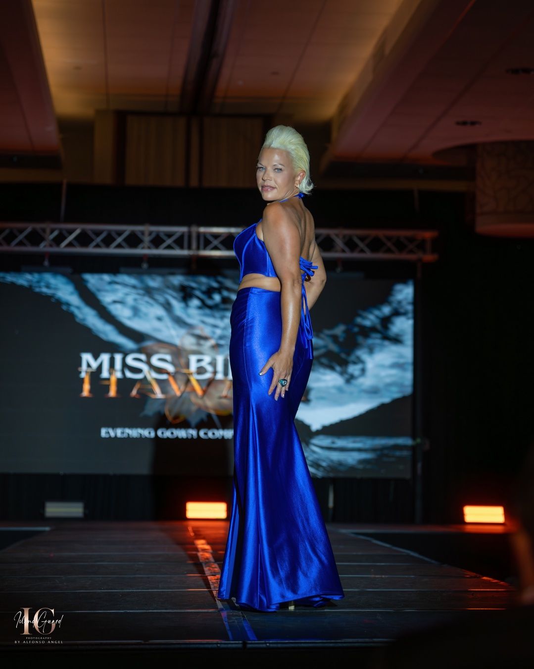 A beauty contestant in a royal blue evening gown poses elegantly on a runway stage with illuminated signage in the background.