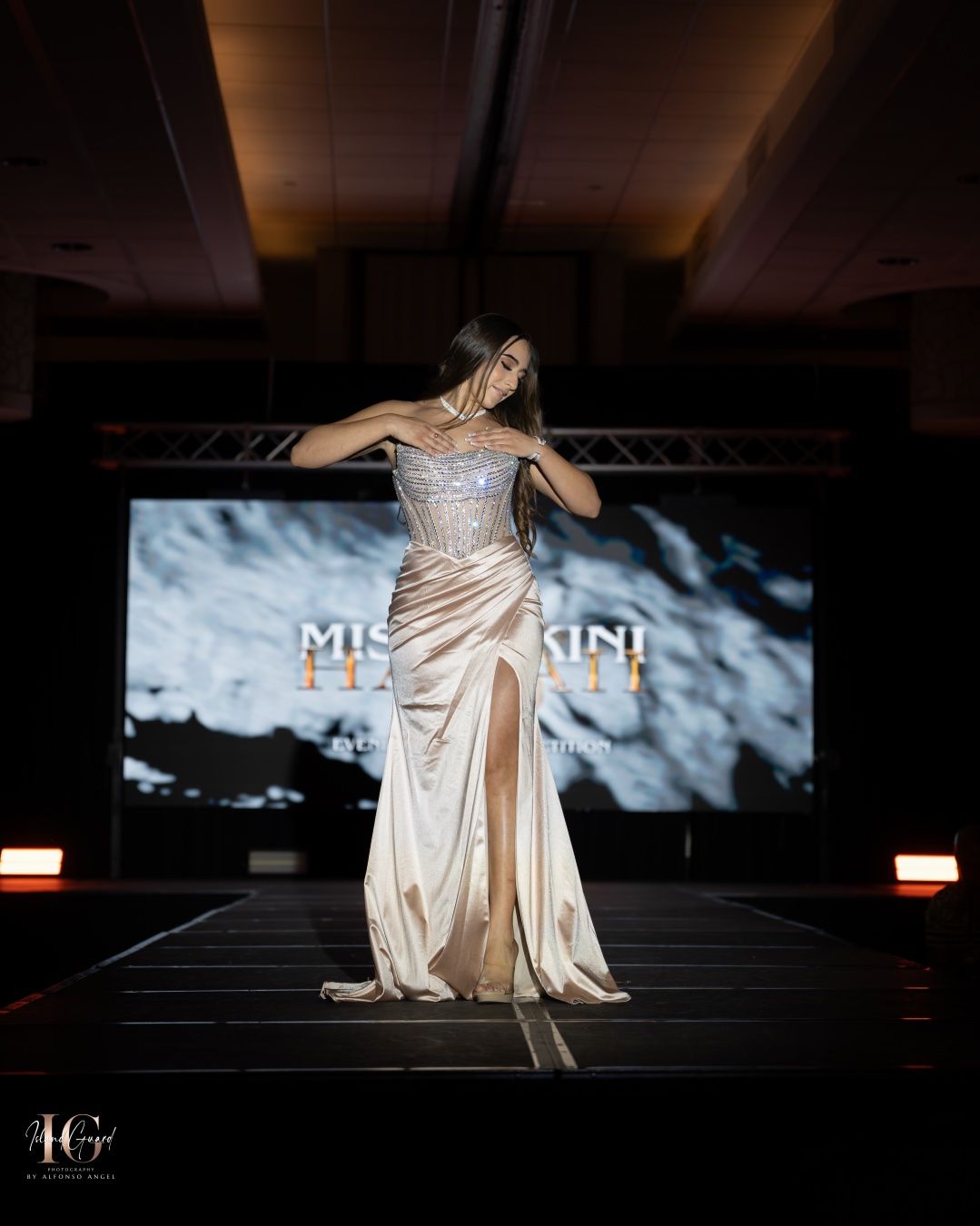 Model showcases elegant champagne silk evening gown with fitted bodice on illuminated fashion runway with display screen.