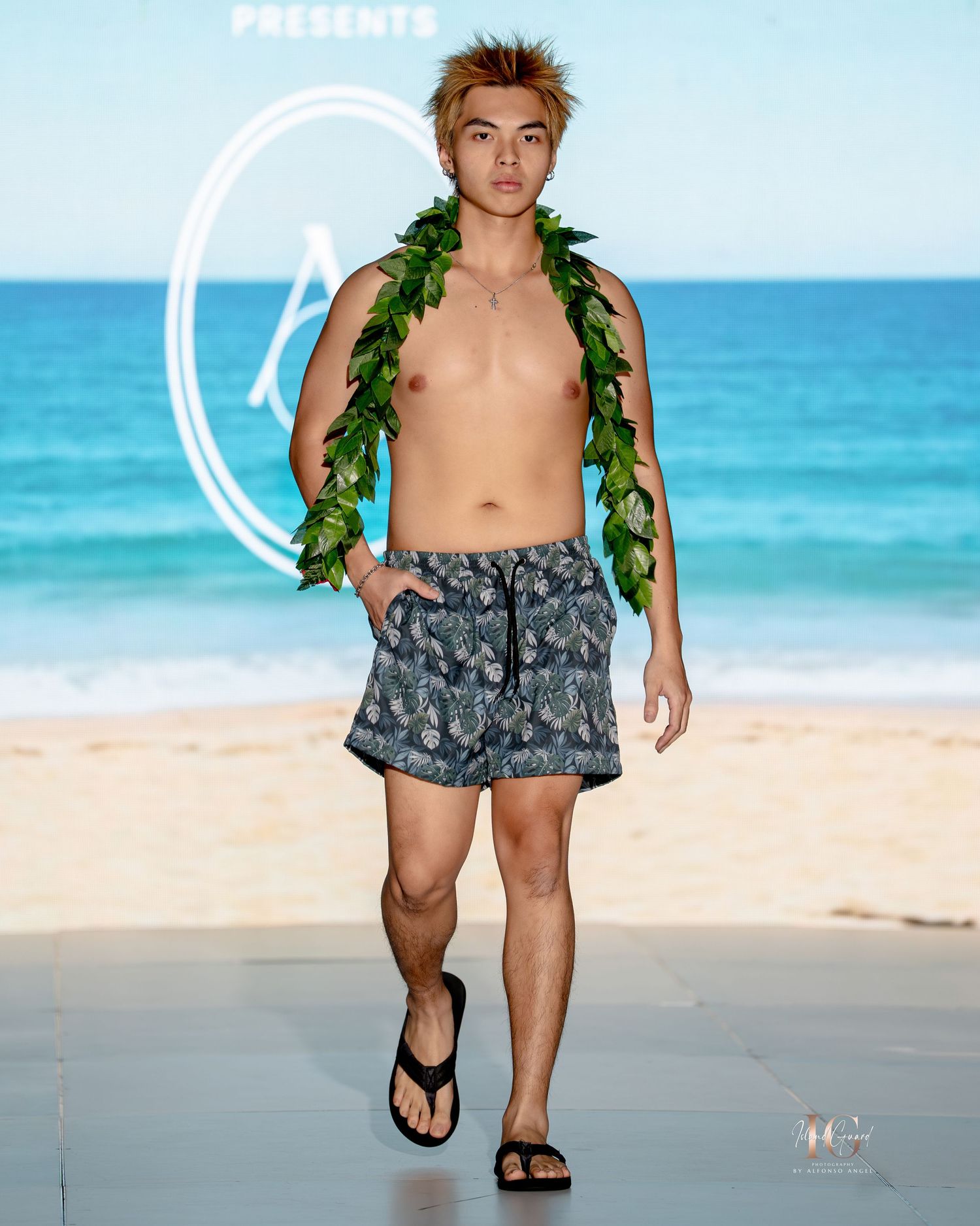 A model walks the runway wearing a green leaf garland and gray patterned swim shorts on a beachfront catwalk.