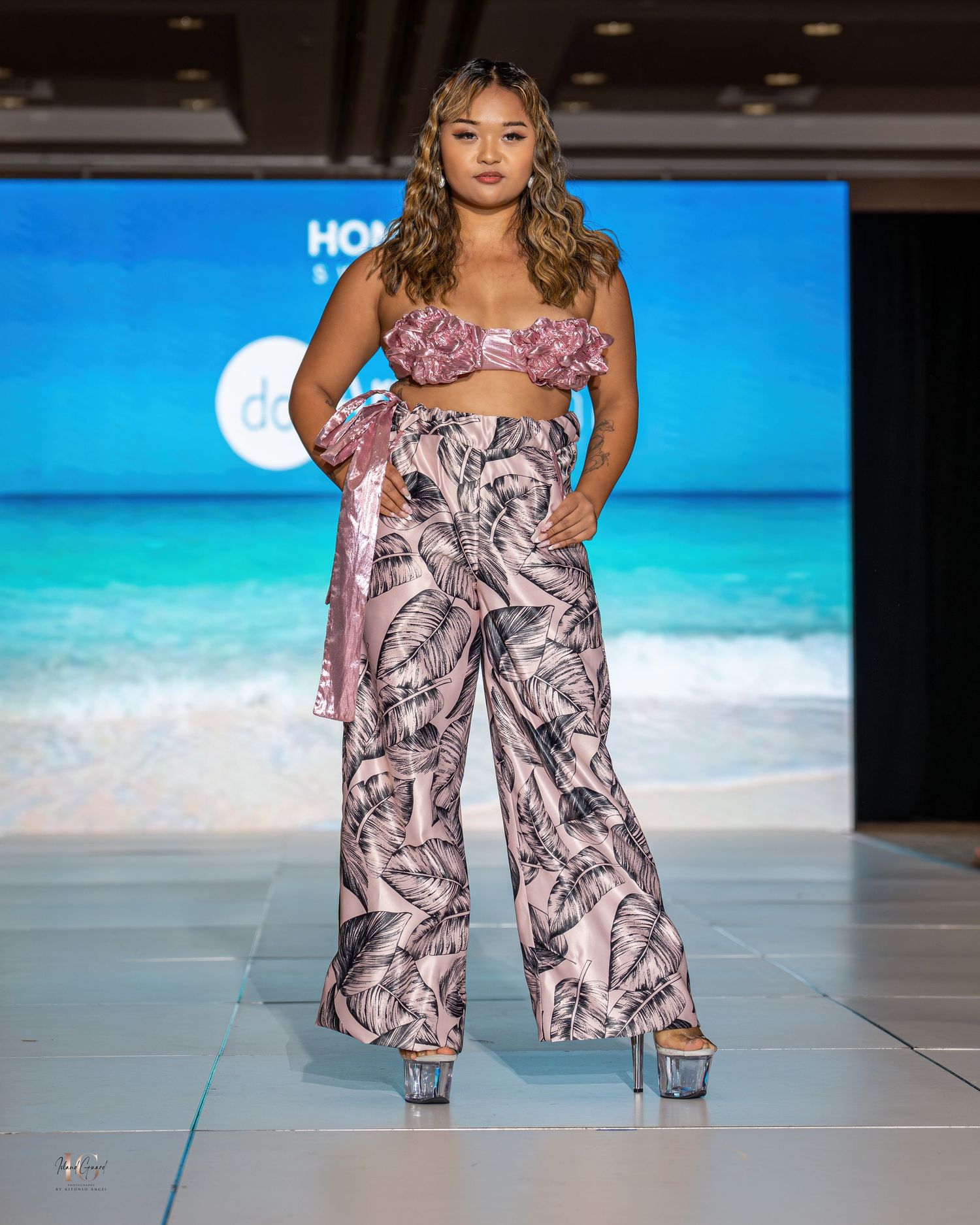 Model showcases floral print wide-leg pants and pink top on beachwear runway with tropical backdrop.