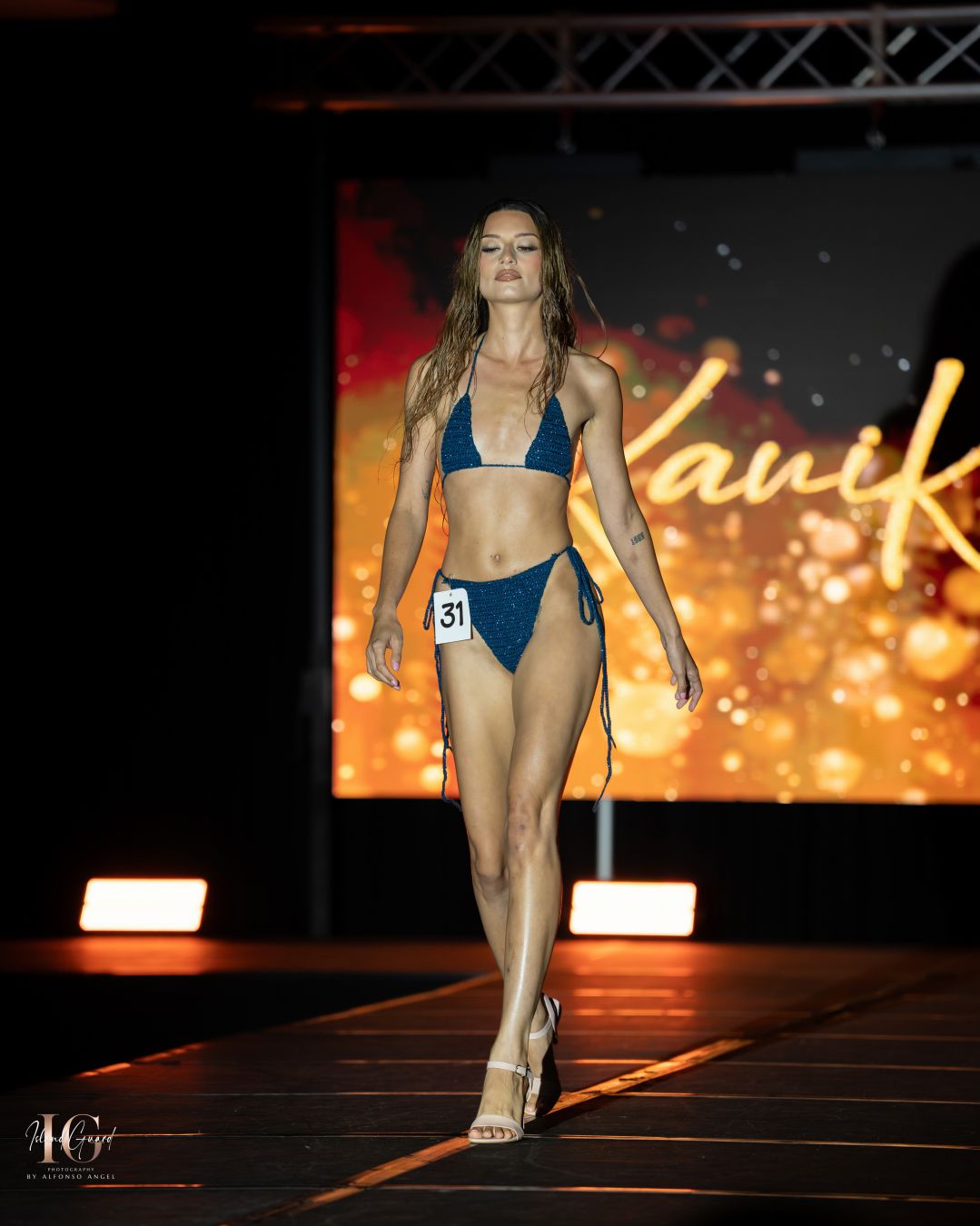 Fashion model presents dark blue swimwear collection on illuminated stage.