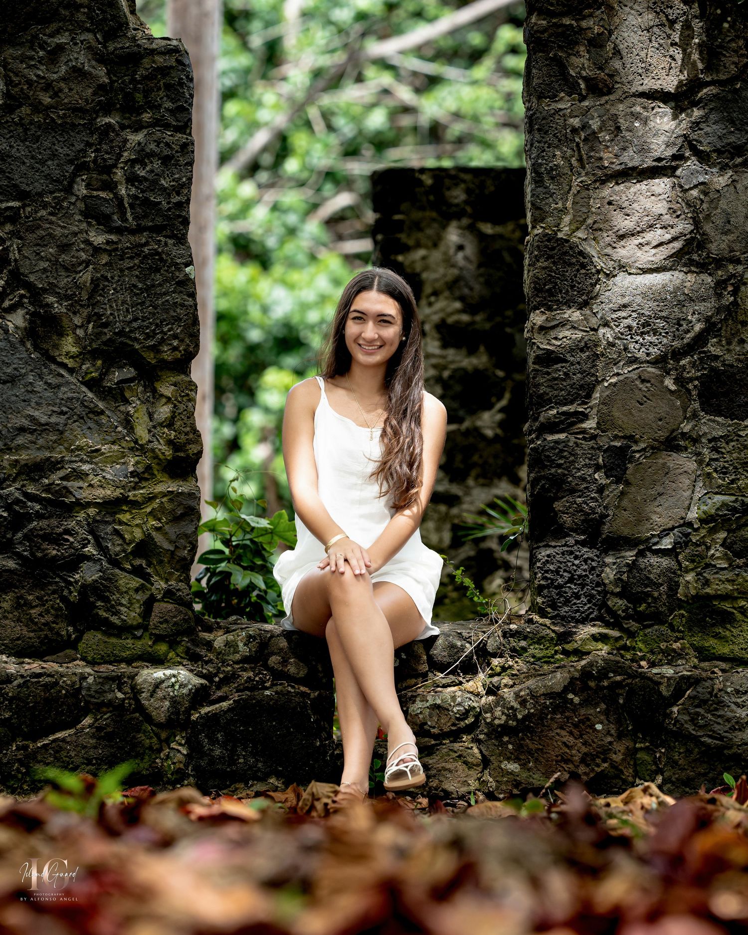 Woman in white dress sitting on old stone ruins surrounded by lush greenery and fallen leaves.