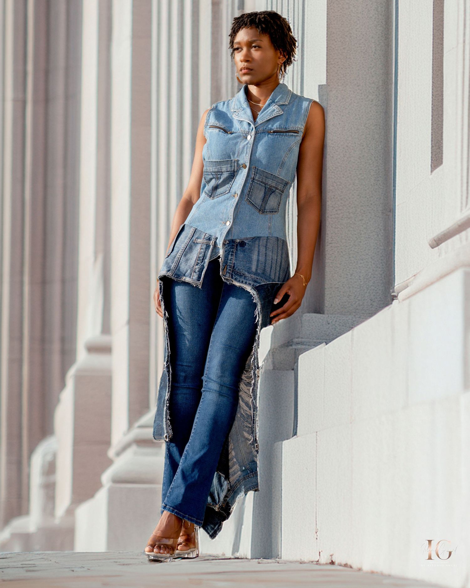Street fashion model wearing a sleeveless denim vest and flared jeans poses against white columns in an urban setting.