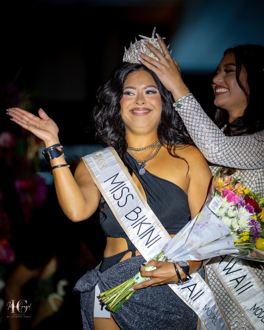 Beauty pageant contestant in a black evening gown holds a bouquet while wearing a sash during an award ceremony.