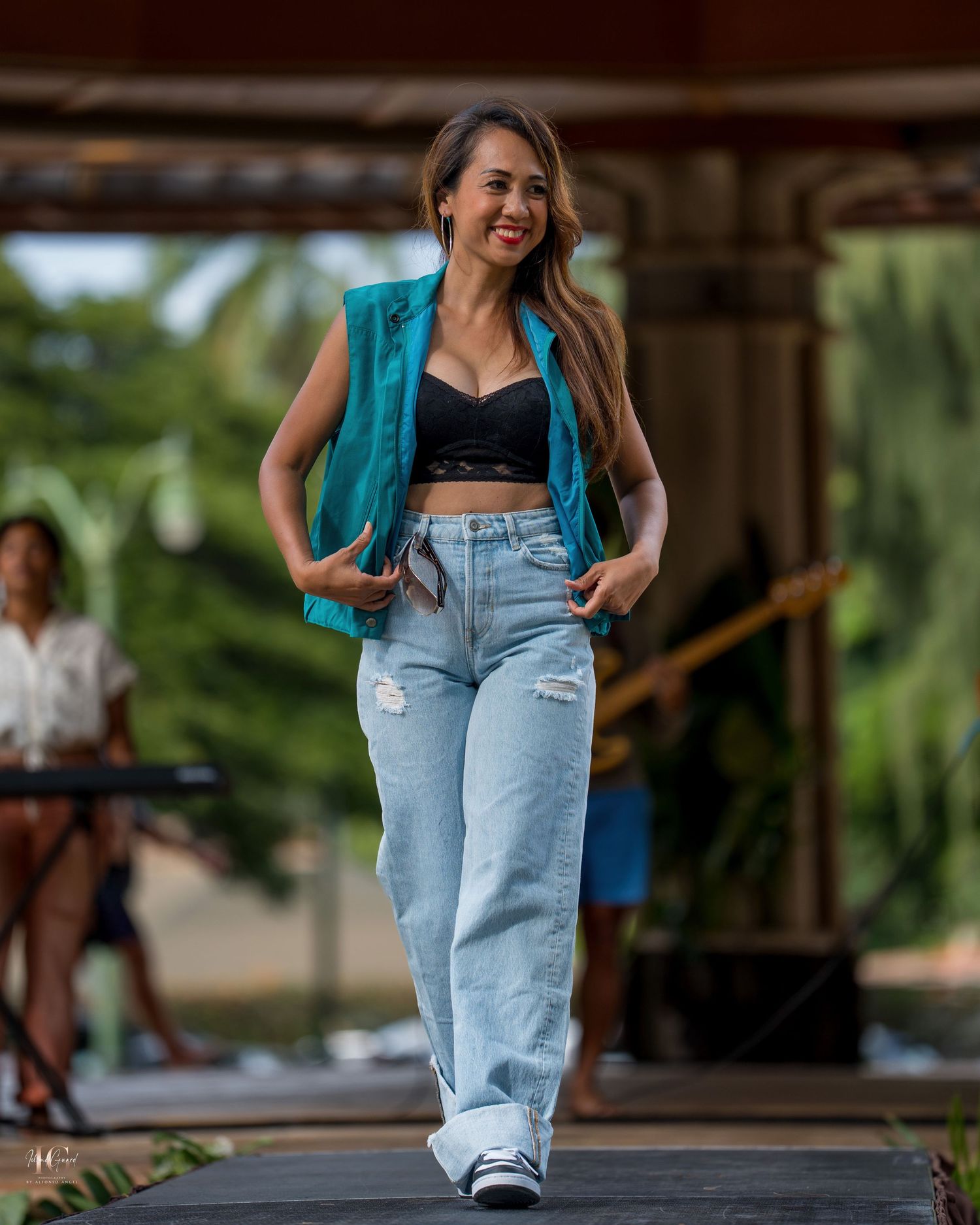Fashionable outfit with turquoise vest, black crop top and baggy light wash jeans worn on outdoor stage or platform.