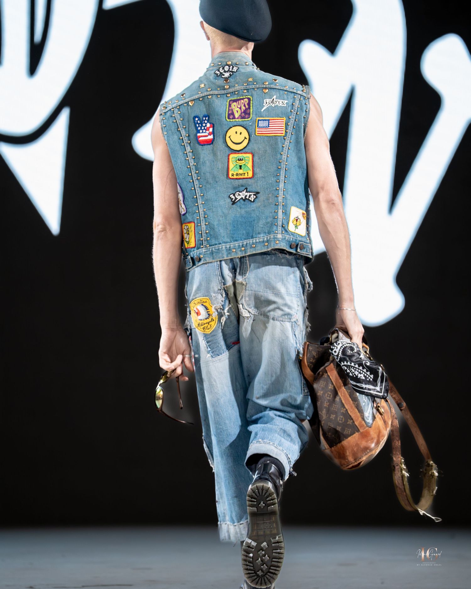 Runway model wearing distressed denim vest with patches and combat boots during fashion show.