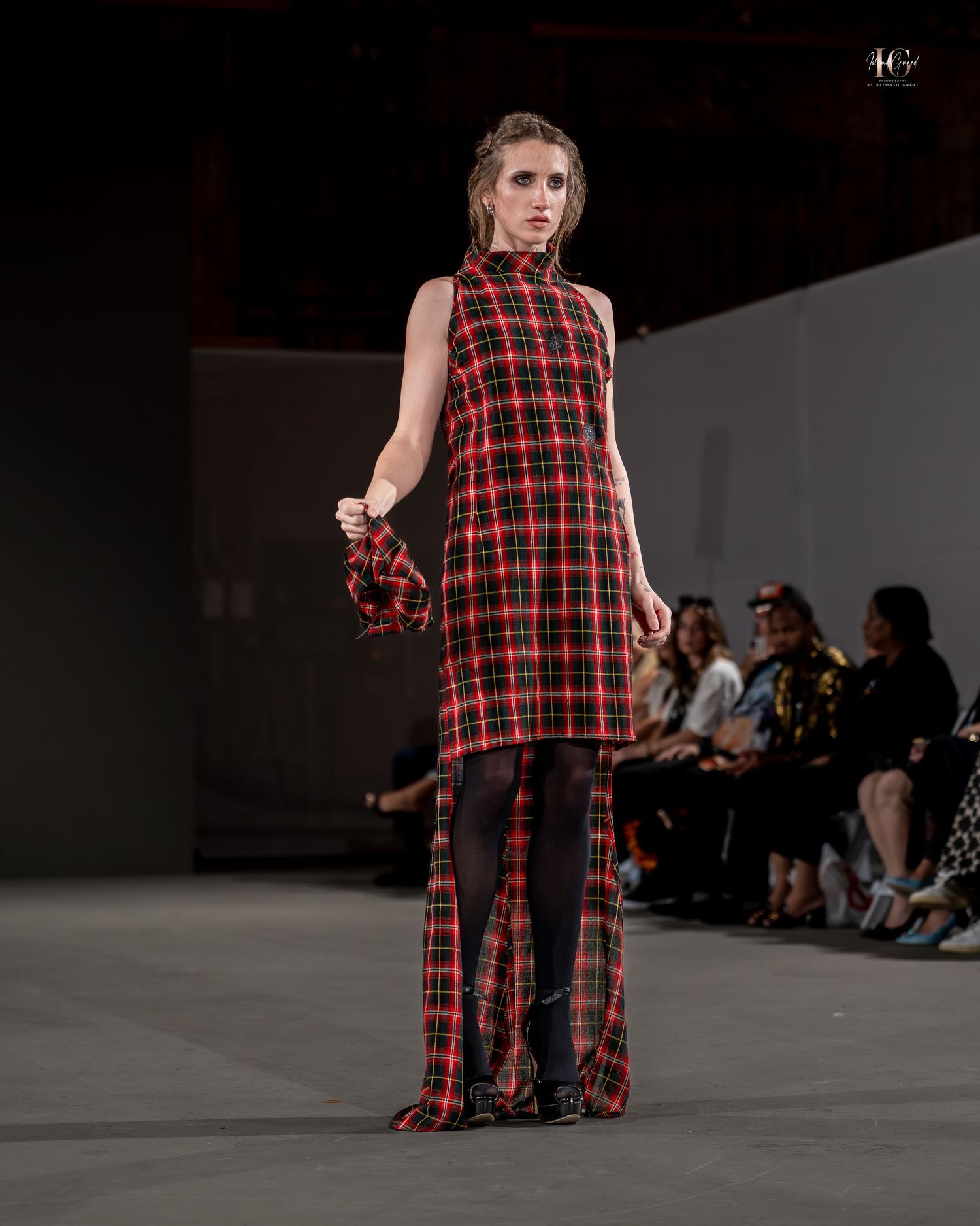 Model walks runway wearing red and black plaid sleeveless dress during fashion show presentation.