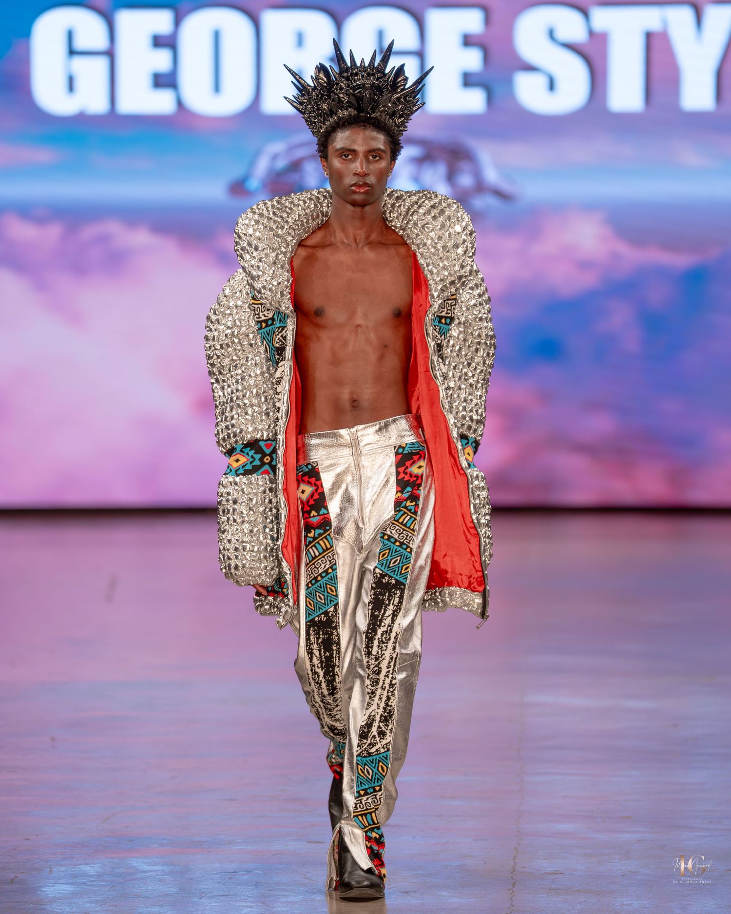 Model walks runway in metallic silver shorts and dramatic winged jacket at fashion show with pink-purple backdrop.