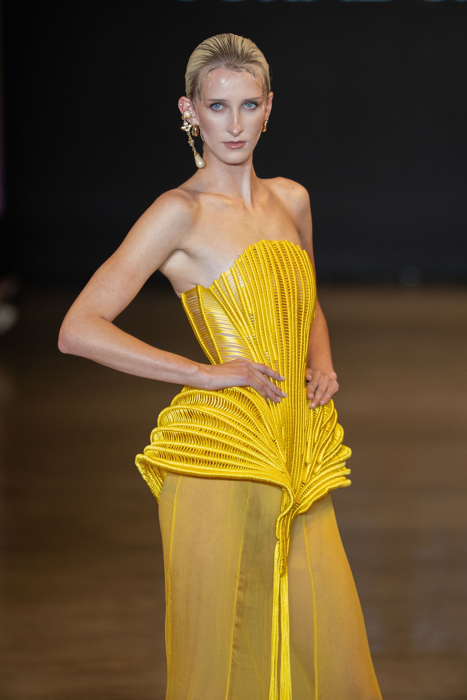 Fashion model wearing a vibrant yellow pleated gown with architectural details on a dark runway.