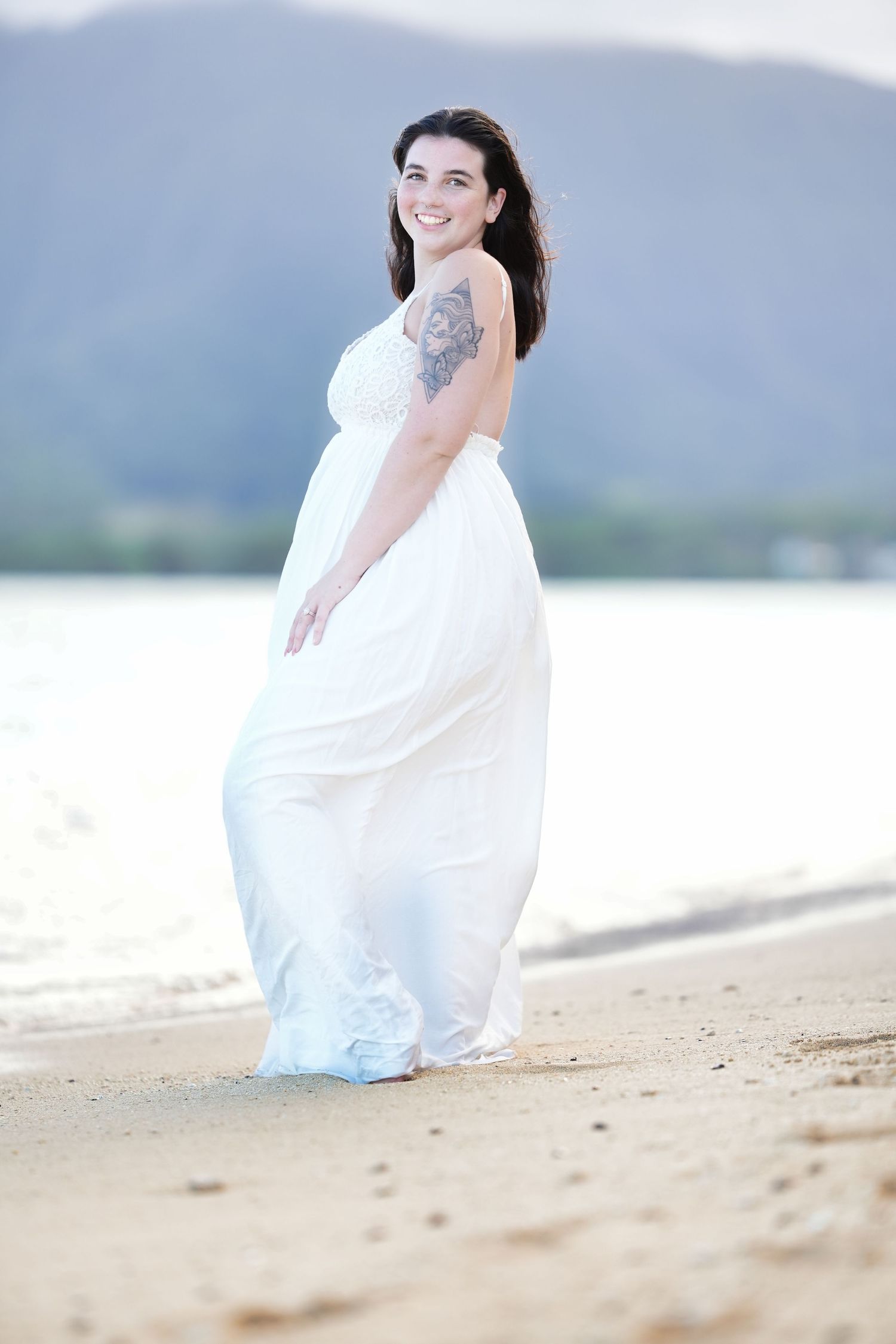 A person wearing a flowing white dress sits gracefully on a sandy beach with mountains in the distant background.