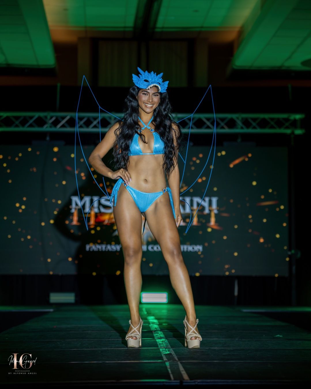 A model in a bright turquoise swimsuit walks confidently down an illuminated runway at a fashion show.
