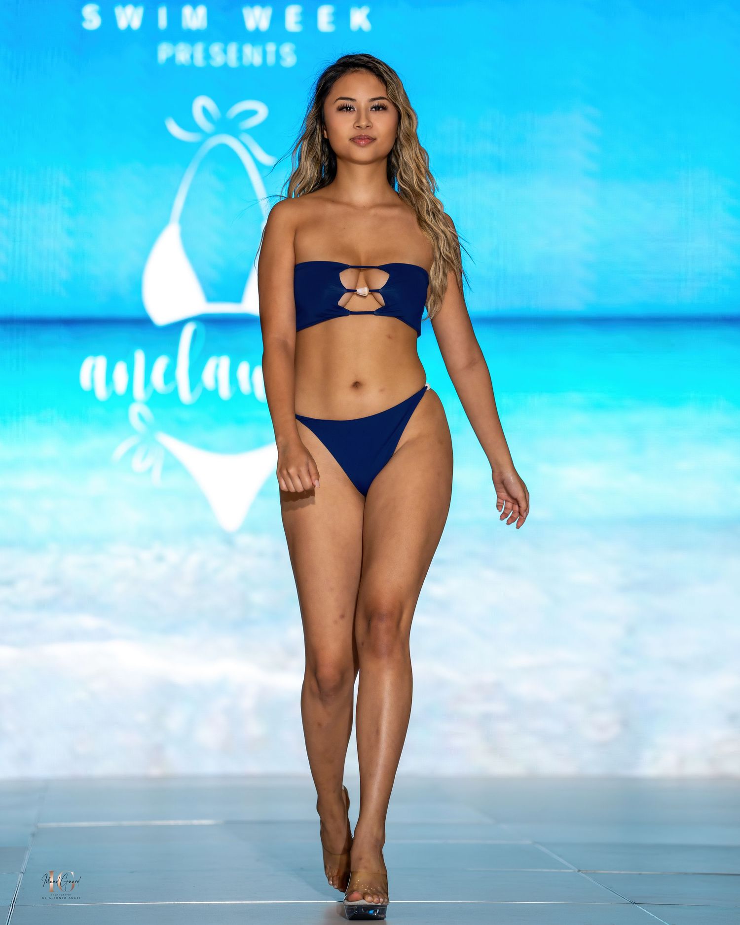 Model walks runway in navy blue two-piece swimsuit at Honolulu Swim Week fashion show.
