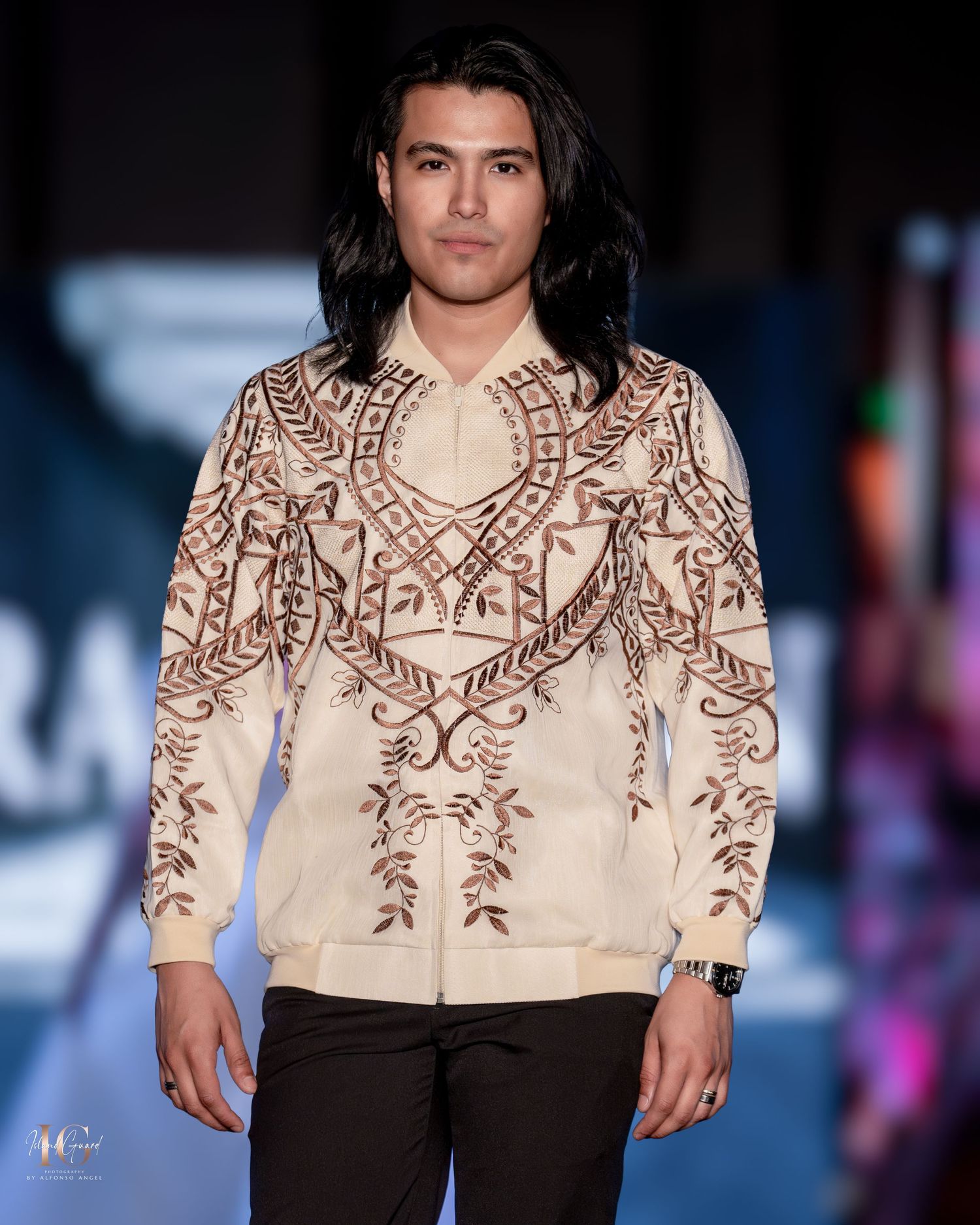 Model presents detailed cream embroidered jacket with dark pants on modern catwalk.