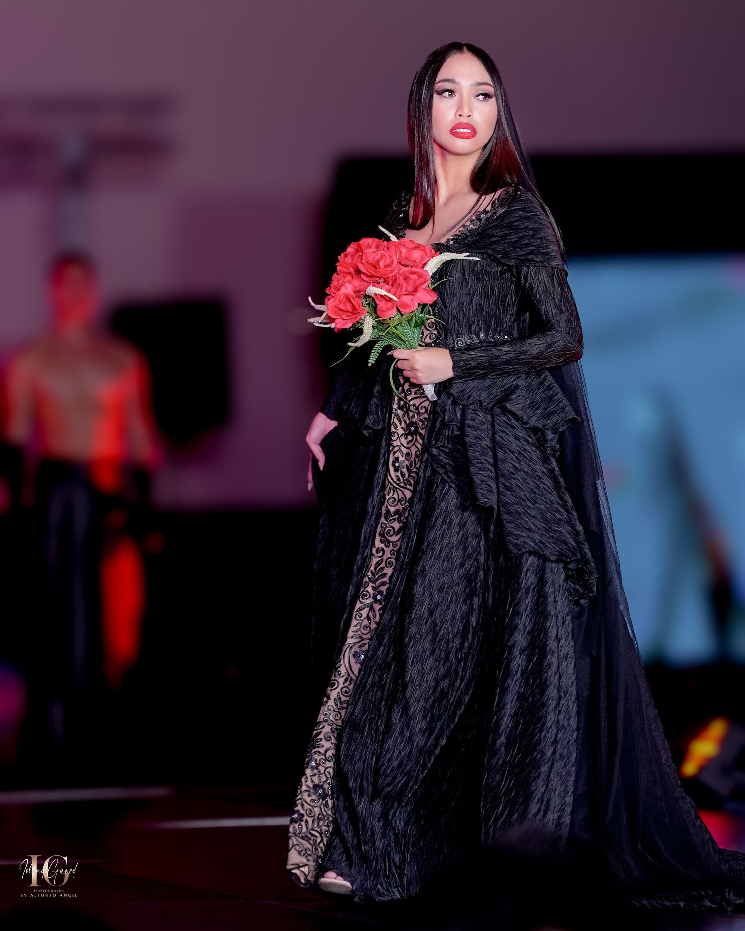 Dramatic black lace gown with red roses displayed at elegant fashion show.