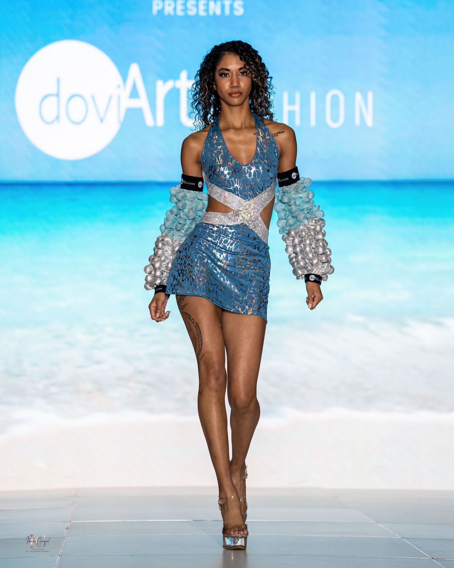 Model presents blue sequined romper with puffy sleeves during beachwear fashion showcase.