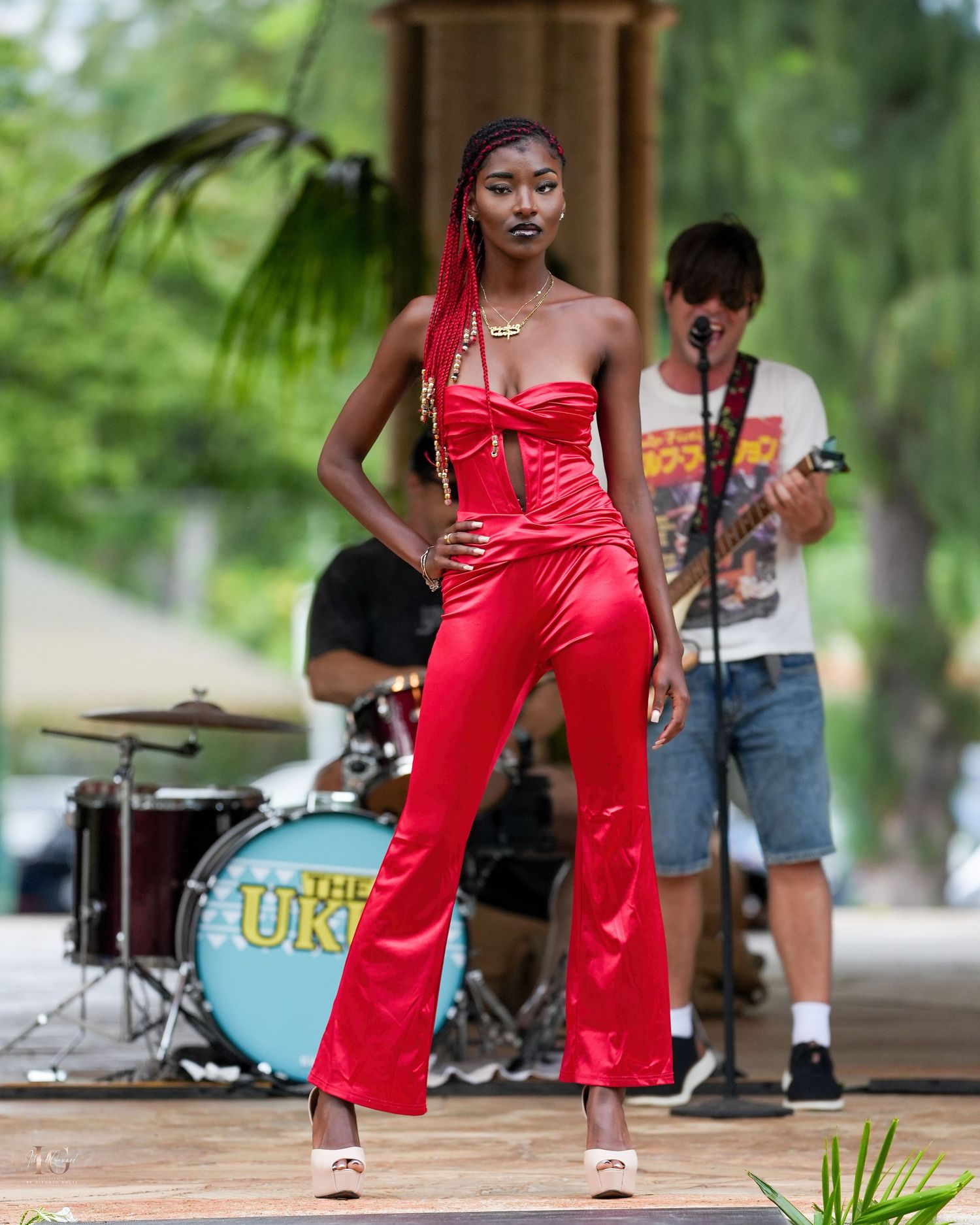 Performer in a vibrant red jumpsuit strikes dynamic dance poses on an outdoor stage with musicians in the background.